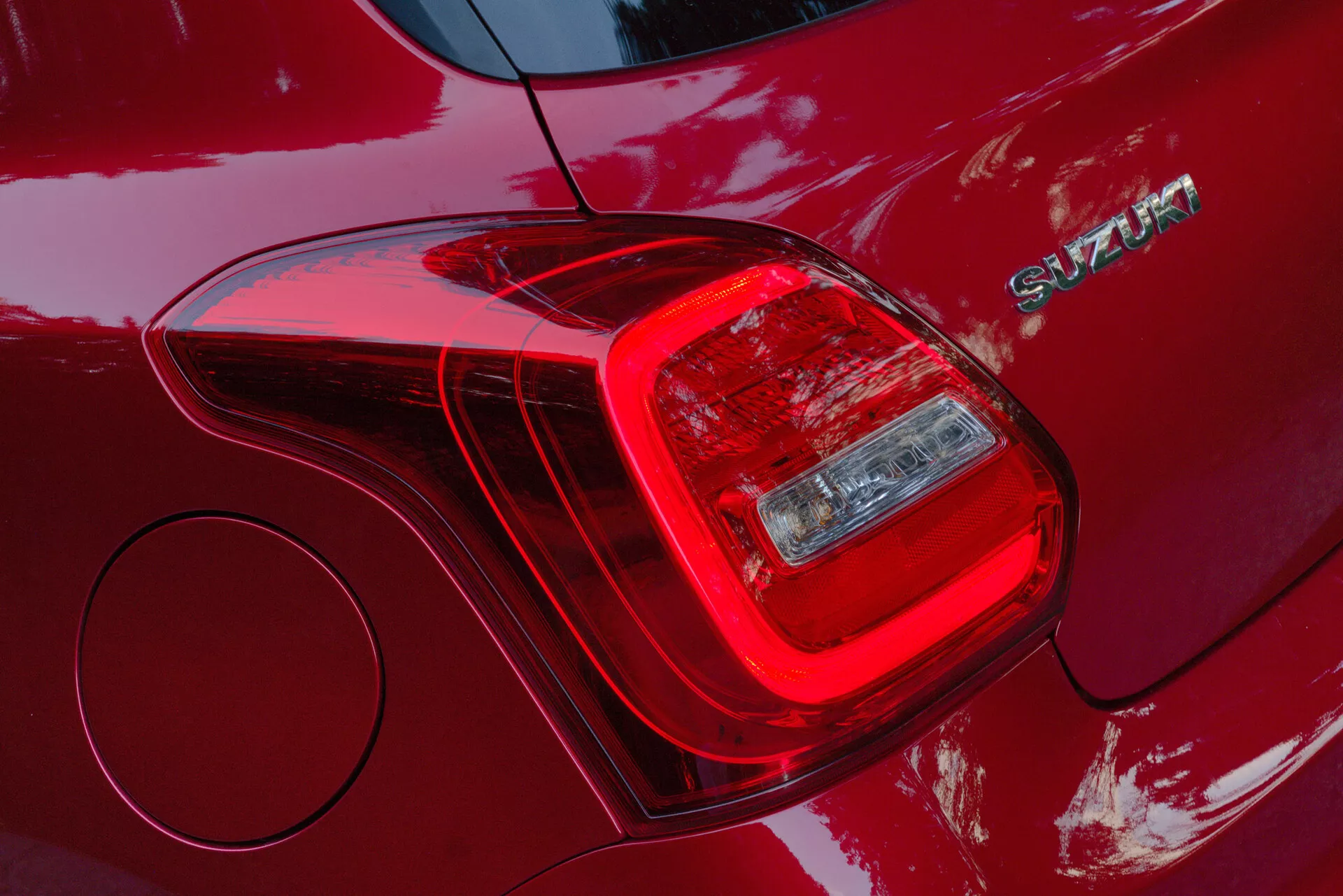 Detailný záber na zadné ľavé svetlo auta Suzuki Swift Sport 2017, zvýrazňujúci segmentovaný dizajn svetelných jednotiek vrátane brzdového svetla a smerovky alebo spätného svetla. Karoséria v tmavo červenej farbe s lesklým povrchom odráža svetlo a okolie. Vedľa zadného svetla je viditeľný chrómový emblém s nápisom 'SWIFT'. Záber tiež odhaľuje kruhový kryt palivovej nádrže a zdôrazňuje moderný dizajn s aerodynamickými líniami a trvácnymi materiálmi na konštrukciu.