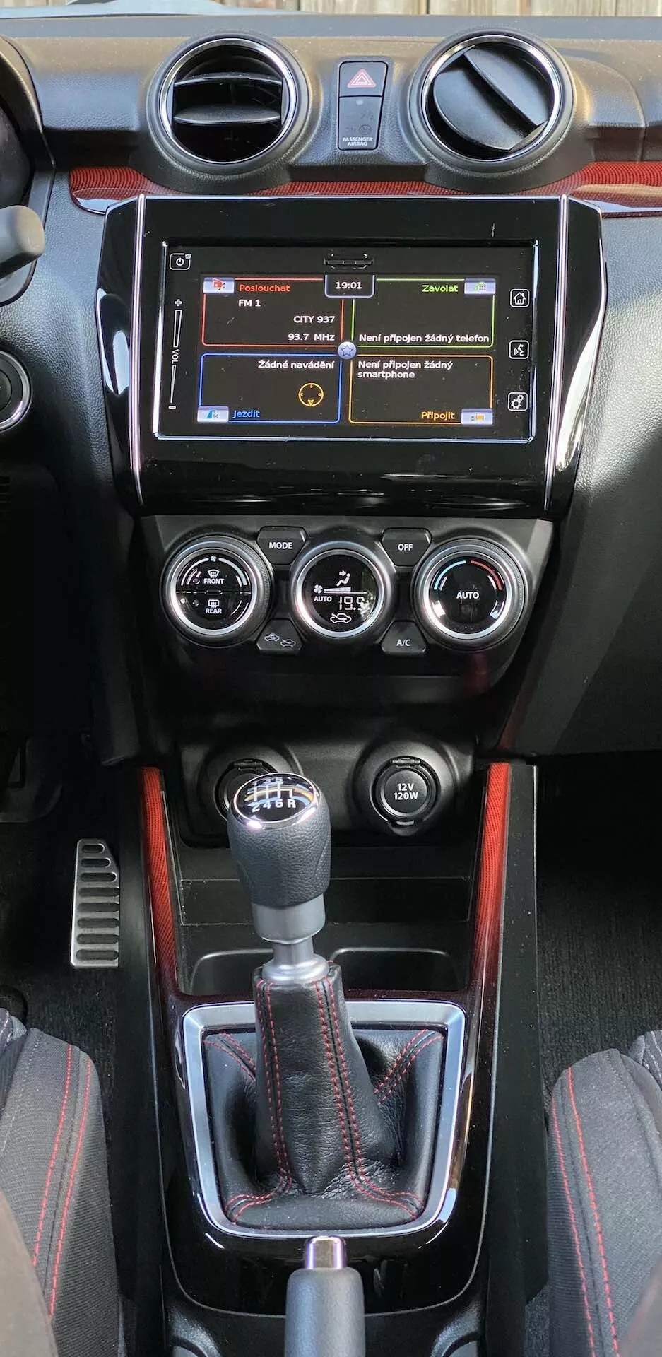 Stredová konzola interiéru vozidla Suzuki Swift Sport 2017, vybavená infotainment systémom s fyzickými tlačidlami, kruhovými vzduchovými ventilačnými otvormi, manuálnym panelom klimatizácie s tromi ovládačmi pre teplotu, rýchlosť ventilátora a rozdelenie vzduchu a športovým otočným gombíkom manuálnej prevodovky. Konzola obsahuje kožený poťah, dekoratívne obloženie pravdepodobne s uhlíkovým vzorom a športovú červenú líniu. Ďalšie ovládacie prvky pod pákou naznačujú pokročilé asistencné systémy.