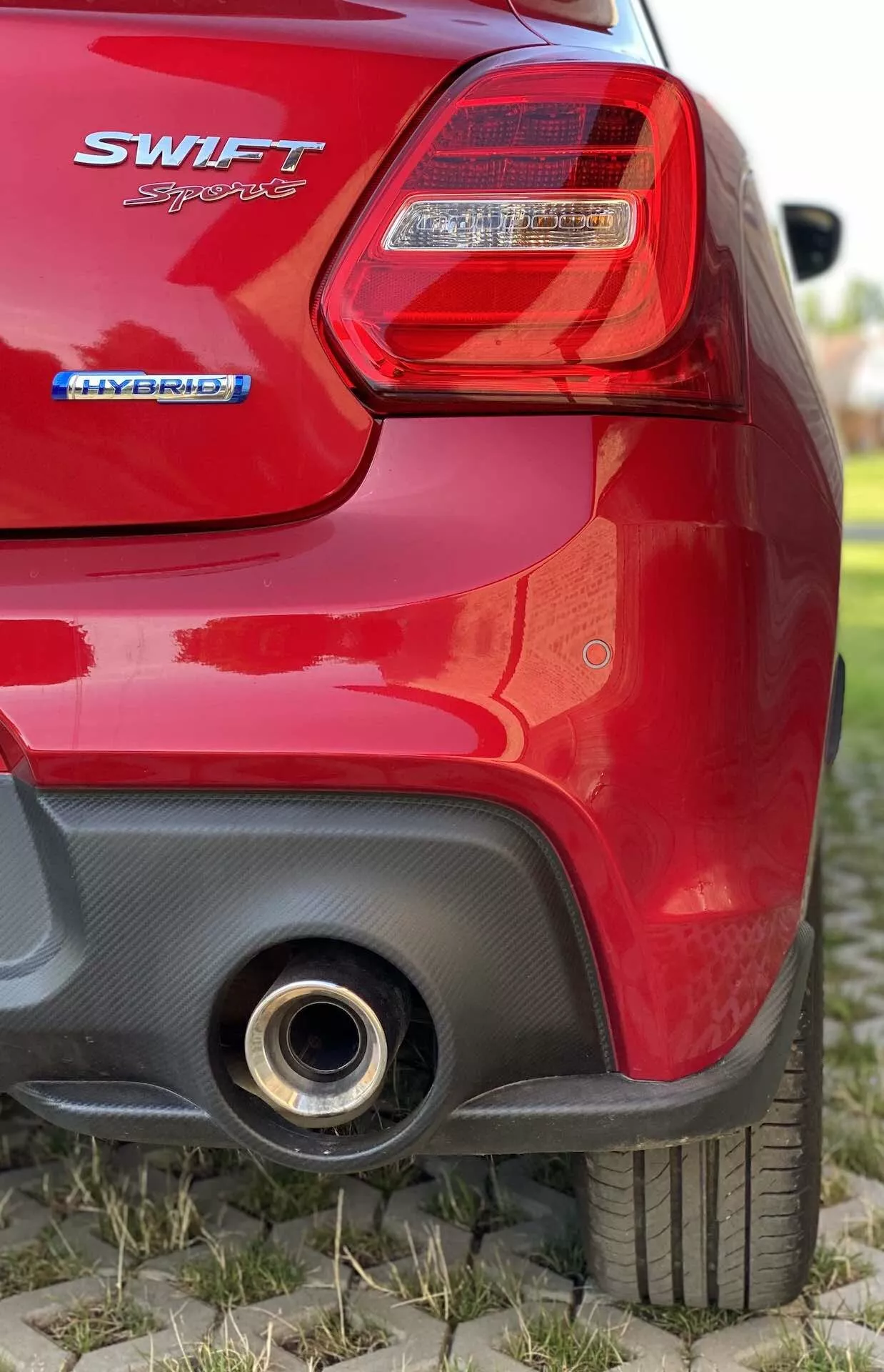 Detailný pohľad na zadnú časť červeného hatchbacku Suzuki Swift 2017 z nízkeho uhla. Obrázok sa sústreďuje na pravé zadné svetlo s čírym krytom a červenými svetelnými sekciami, znak 'SWIFT' pod svetlom, pod ním znak 'HYBRID' indikujúci hybridný pohon, čiastočne rozostrený zadný kufor, pravé zadné koleso s viditeľným vzorom behúňa, čiernu kontrastnú časť na zadnom nárazníku s možným aerodynamickým prívodom a okrúhlu kovovú výfukovú rúrku v pravom dolnom rohu, ktorá odráža svetlo.