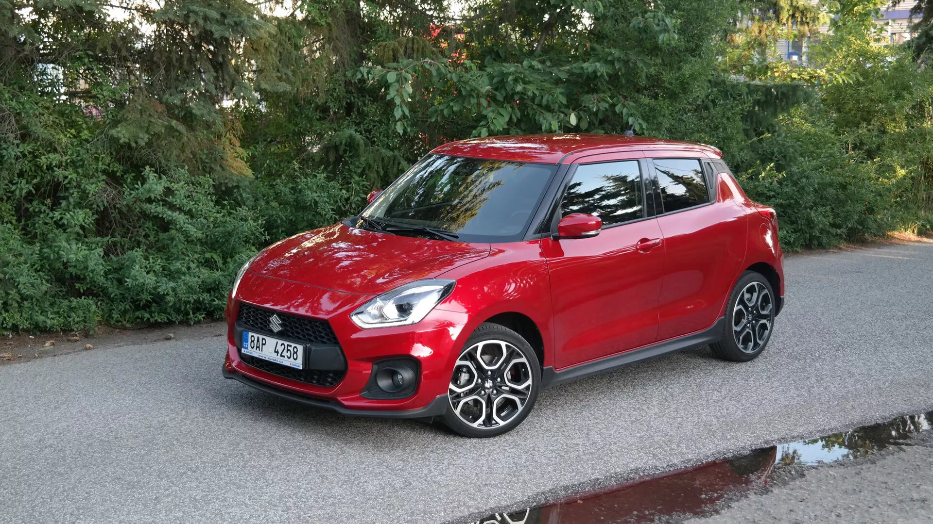 Obrázok červeného hatchbacku Suzuki Swift Sport 2017, vyfotografovaného z trojštvrťového predného uhla, ukazujúceho elegantné svetlomety, výraznú prednú masku s horizontálnymi líniami, hladkú kapotu, jasné čelné sklo, vodičovu stranu vrátane zrkadla farby karosérie, viaclúčové hliníkové kolesá a strešné spojlery proti vydláždenej ceste s prírodnými kulisami.