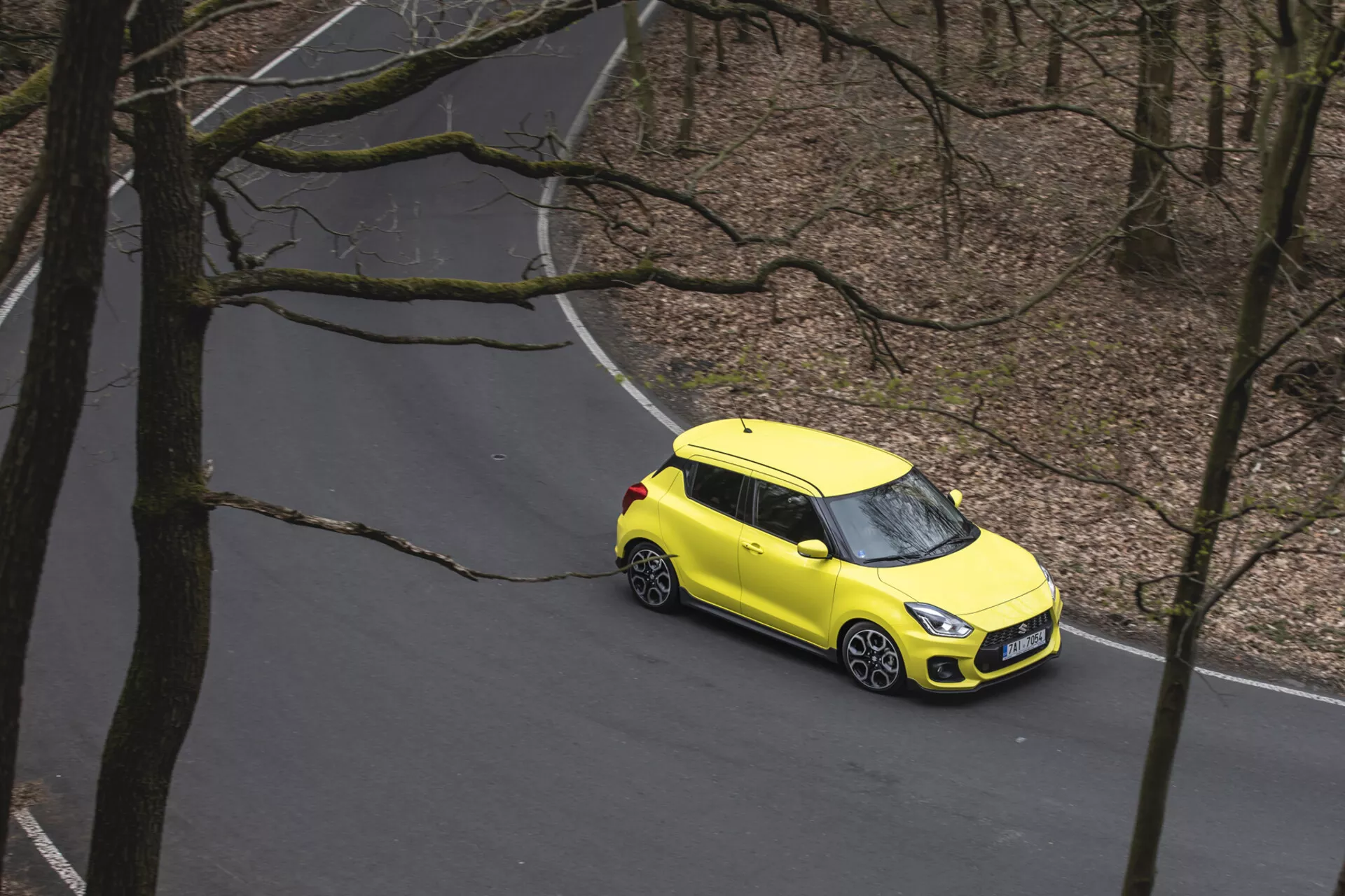 Letecký pohľad na jasne žltý Suzuki Swift 1995 s dvojfarebnou farebnou schémou, cestujúci po serpentíne cez les, predvádzajúci zliatinové kolesá, moderné LED predné svetlomety a kompaktný dizajn hatchbacku.