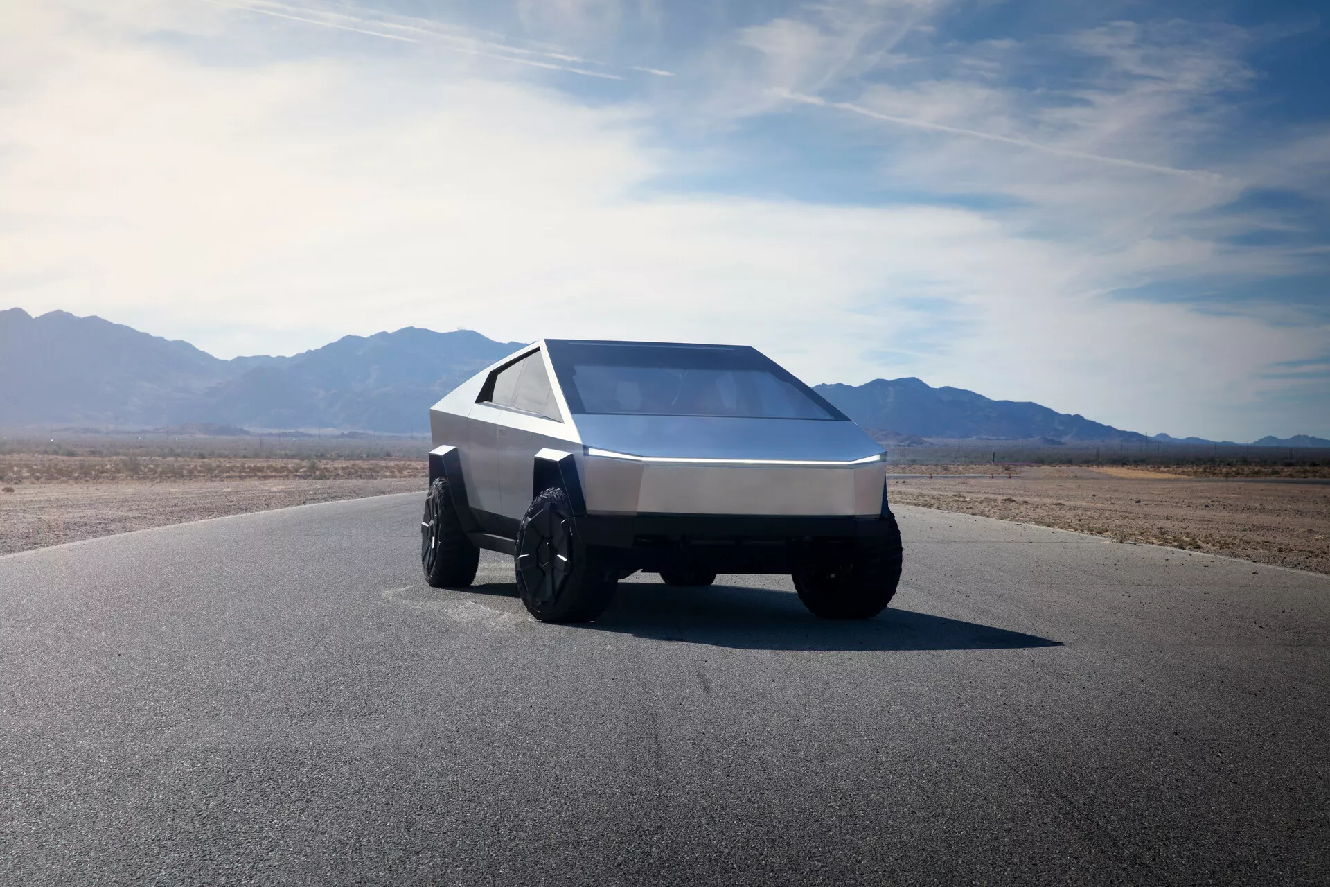 Tesla Cybertruck 2021 (1. generácia) vyfotografovaná z nízkeho predného trojštvrtinového uhla, predstavujúca futuristický, hranatý dizajn. Tento pohľad zachytáva ľavú prednú stranu vozidla, zdôrazňujúc jeho klinovitý tvar s ostrými čiarami a rovnými povrchmi. Vozidlo je vybavené veľkými, robustnými pneumatikami vhodnými pre terénnu jazdu a obsahuje jediný, nepretržitý svetelný pás cez šírku prednej časti, ktorý naznačuje moderné LED alebo laserové osvetlenie. Strmo sklonené, rozľahlé čelné sklo a ostro zužujúce sa bočné okná prispievajú k jeho geometrickému vzhľadu. Vozidlo je vyrobené z brúseného kovu alebo kompozitného materiálu, čo naznačuje trvanlivosť a robustnosť. Pozadie zahŕňa rozsiahlu čistú oblohu a hornatý terén, naznačujúc suché, púštne prostredie.