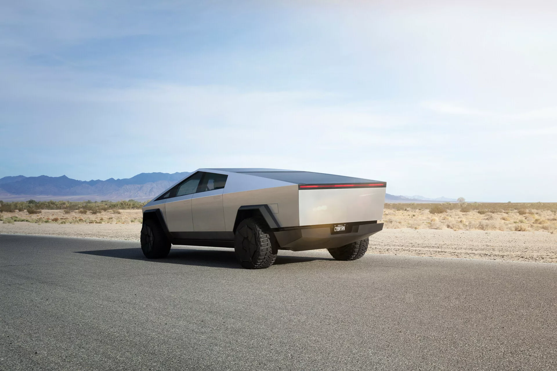 Futuristický Tesla Cybertruck 2021 na rovnej asfaltovej ceste zachytený z uhla troch štvrtín zozadu, ukazuje jeho charakteristický hranatý dizajn, kovovú povrchovú úpravu, veľké terénne pneumatiky a absenciu tradičných zrkadiel a kľučiek dverí, čo naznačuje pokročilé automatizované mechanizmy.