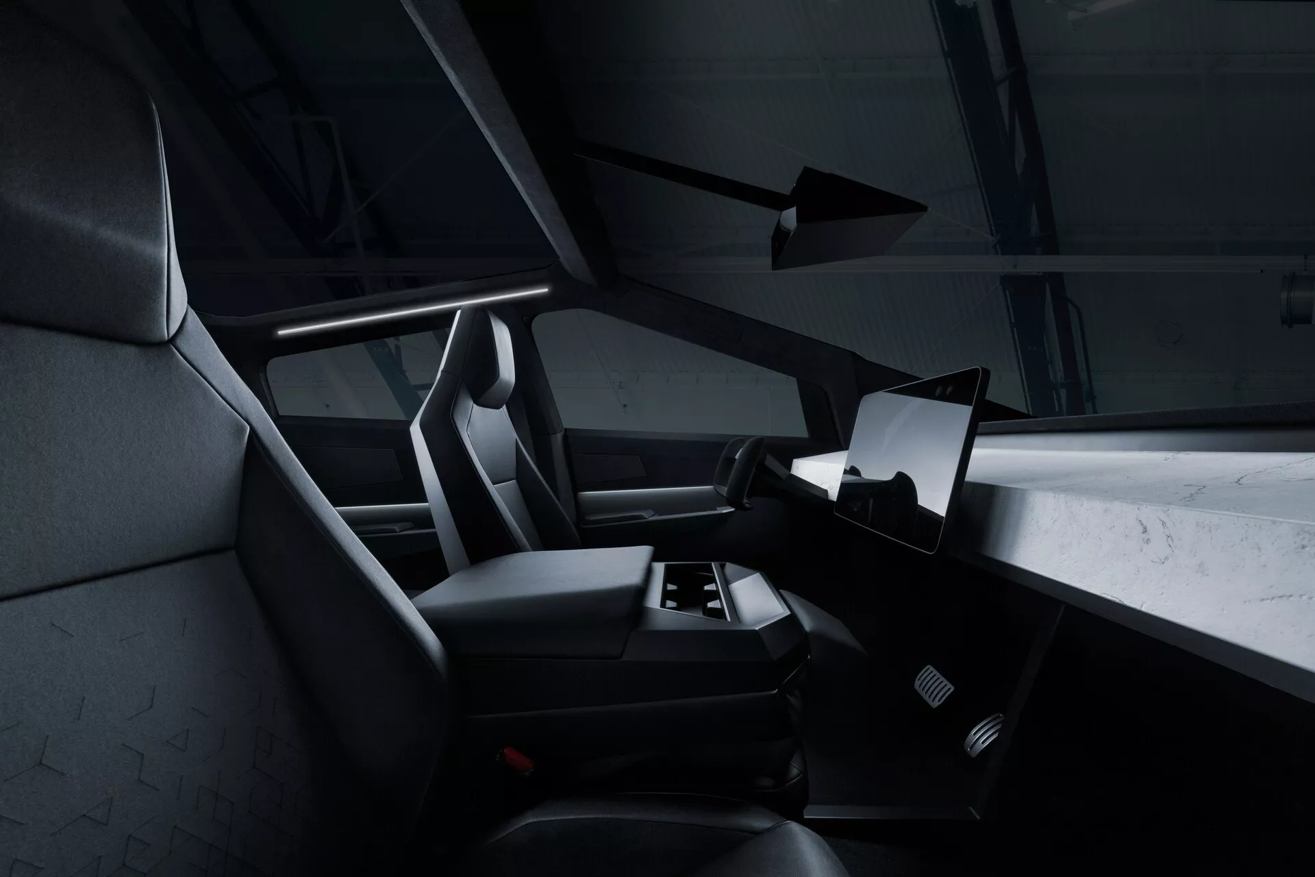 Interiér vozidla Tesla Cybertruck 2021 (1. generácia) z pohľadu zo zadných sedadiel, ukazujúci pravú stranu kabíny s veľkým horizontálne orientovaným dotykovým displejom dominujúcim v strede palubnej dosky. Časť volantu je viditeľná, ale väčšinou zakrytá. Predné sedadlá pre spolujazdca a vodiča sú so zabudovanými opierkami hlavy a textúrovaným čalúnením, pravdepodobne z látky alebo syntetickej kože. V priestore podlahy je viditeľná rohož, a nie sú viditeľné žiadne manuálne ovládacie prvky, čo poukazuje na minimalistický, digitálny prístup. Tmavá farebná paleta a jemné akcentové osvetlenie dodávajú moderný, luxusný vzhľad.