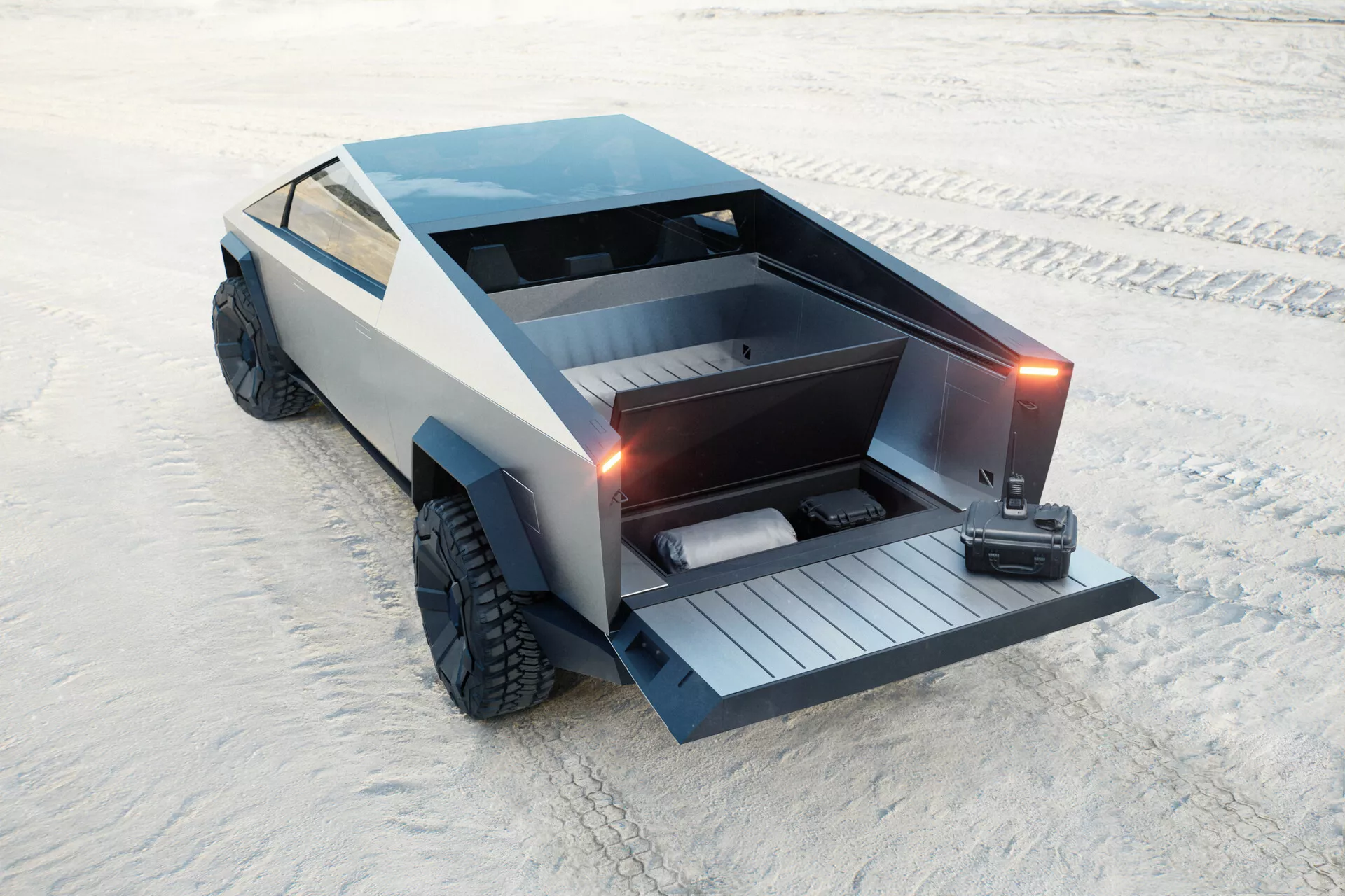 Futuristický Tesla Cybertruck 2021 (1. generácia) zaparkovaný na piesočnatom povrchu, zachytený z vyvýšeného zadného trojštvrťového pohľadu. Uhlový, polygonálny dizajn s plochou korbou a motorizovaným sklopeným zadným dverami, ktoré odhaľujú vstavanú rampu. Vozidlo obsahuje aj uzamykateľný kryt tonneau, veľké terénne pneumatiky a matný sivý povrch, ktorý prispieva k jeho minimalistickej, priemyselnej estetike. Nie sú viditeľné tradičné kľučky dvier ani bočné zrkadlá, čo naznačuje zasúvacie kľučky a zrkadlá na báze kamier. Predpokladom je elektrické s dôrazom na praktickosť a terénne schopnosti.