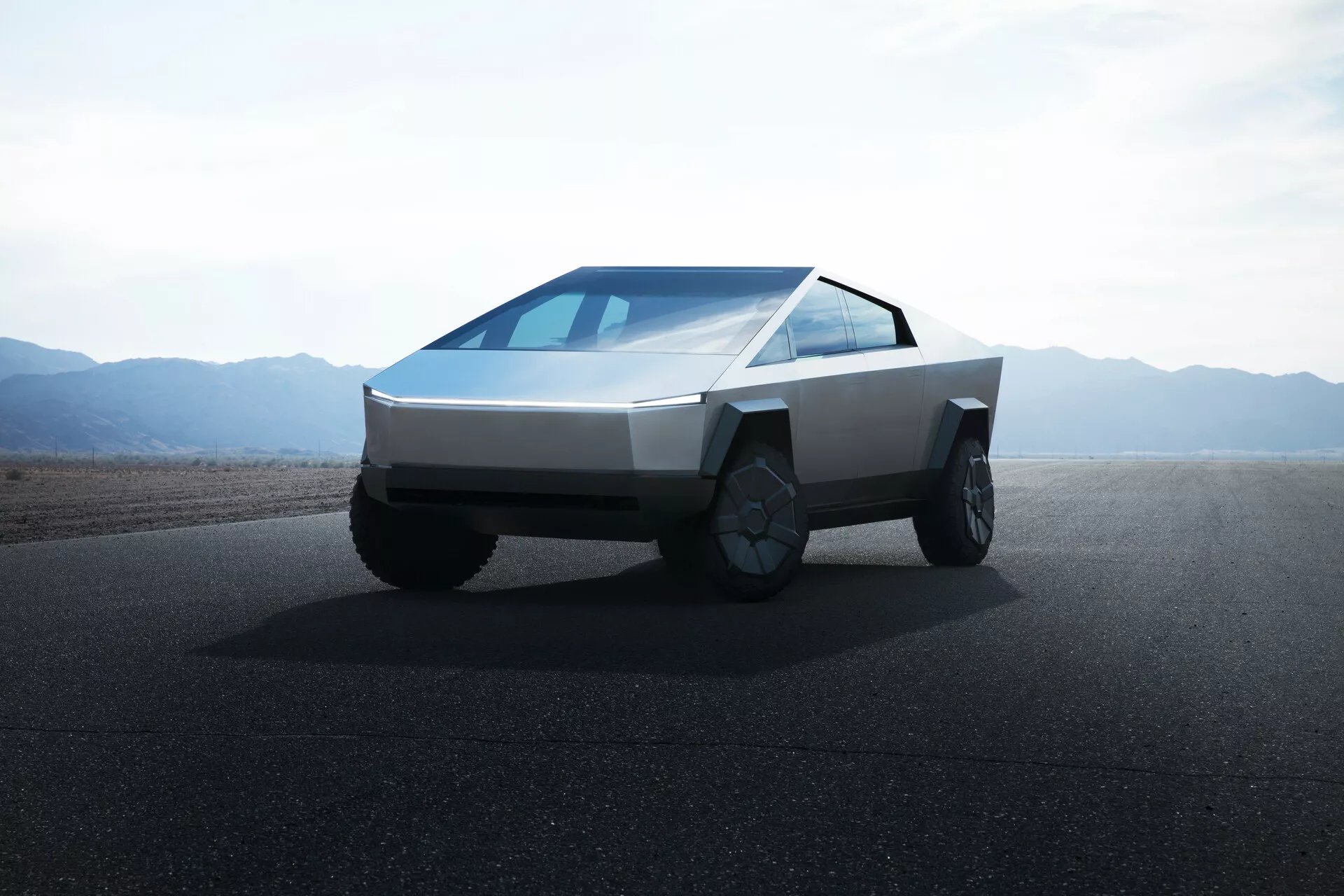 Obrázok Tesla Cybertruck 2021 (1. generácia) na ceste s rovinatým terénom alebo púštnymi horami v pozadí za denného svetla a jasnej oblohy. Fotografia je urobená z nízkeho uhla, čím poskytuje záber na prednú štvrtinu vozidla a zdôrazňuje jeho futuristický, hranatý dizajn. Vozidlo má klinovitý predok, telo z plochých panelov, tenké horizontálne svetlomety, kontinuálne strmý čelného skla, žiadne viditeľné bočné zrkadlá, veľké kolesá schopné terénnej prevádzky so zložitými ráfikmi a chýba predná mriežka, ktoré naznačuje elektrický pohon.