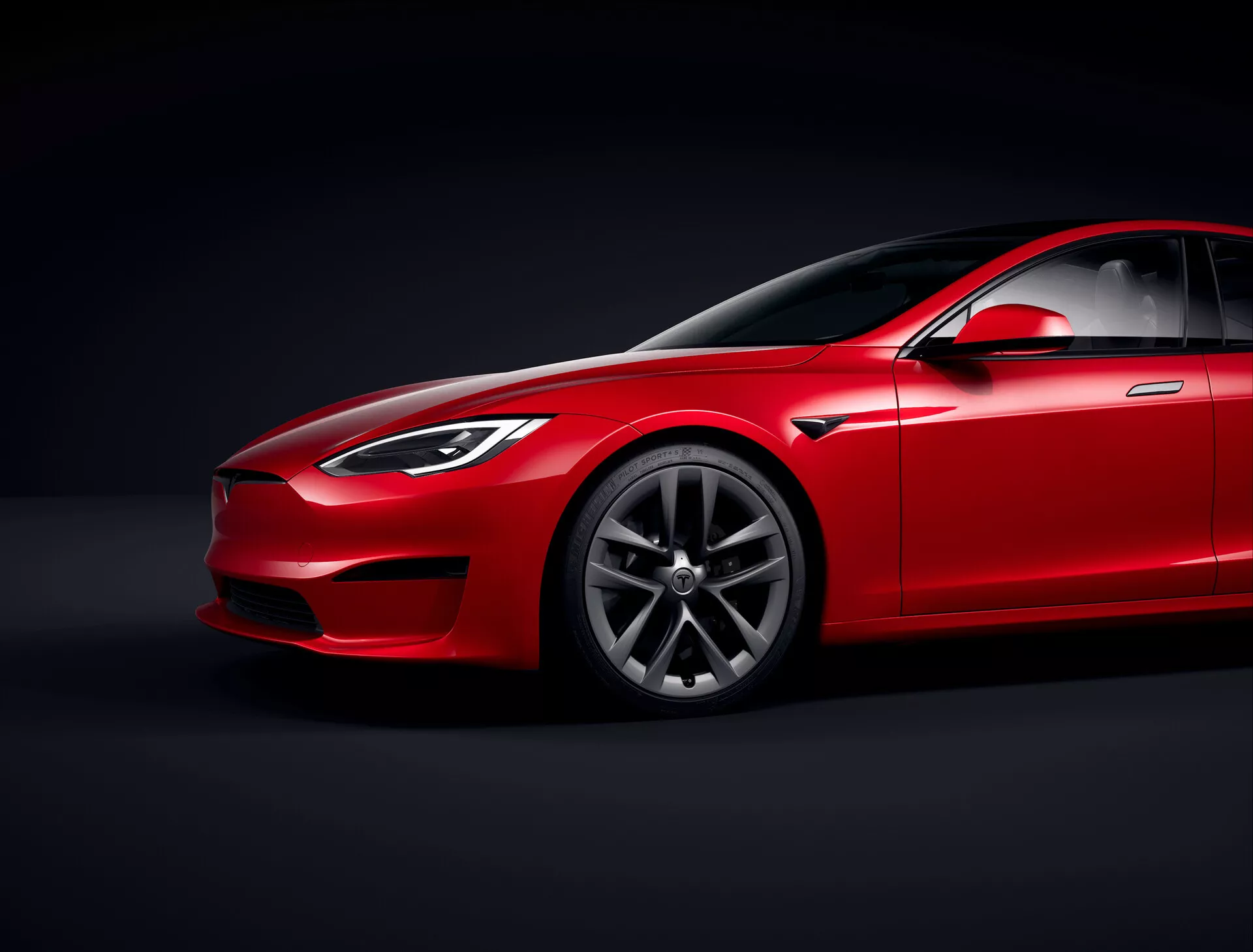 Tesla Model S 2016 (facelift prvej generácie) červený sedan v štúdiu s riadeným osvetlením a čiernym pozadím, nízky uhol pohľadu zdôrazňujúci aerodynamický profil a svetlú výšku, trojštvrtinový predný pohľad ukazujúci stranu vodiča, svetlomety, mriežku, časť kapoty, dizajn ráfikov s prémiovými viaclúčovými vzormi, čelné sklo s tónovaným sklom, sklonenú líniu strechy, bočné zrkadlo, kľučky dverí a líniu okna hladko sa integrujúcu do štíhleho dizajnu, zdôrazňujúcu povrchovú úpravu laku, kontúrne čiary a tvarované panely naznačujúce luxus a výkon, zamerané osvetlenie zobrazujúce detaily dizajnu.
