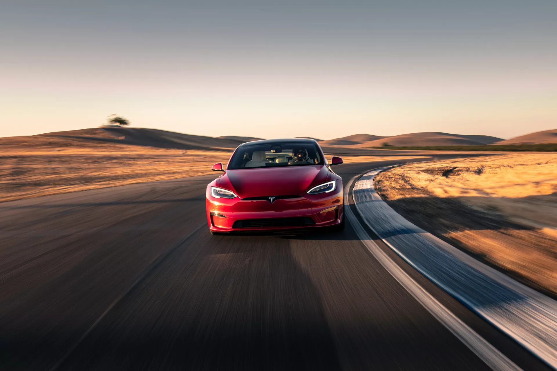 Obrázok červeného sedanu Tesla Model S 2016 (prvá generácia facelift) jazdiaceho po zakrivenej ceste, zachyteného z trojštvrťového predného uhla. Fotografia zvýrazňuje ľavú prednú časť vozidla, vrátane loga, svetlometov a časti kapoty, ktorá sa tiahne až k zadnej časti. Chýbajúca tradičná mriežka chladiča poukazuje na elektrický charakter vozidla. Pozadie je rozmazané efektom pohybu, čo naznačuje rýchlosť, pravdepodobne dosiahnutú technikou panorámovania. Auto je umiestnené blízko k objektívu, čo vyvoláva dojem stlačenej perspektívy a odráža moderné technologické pokroky, ako je pokročilý pohonný systém, pravdepodobne autopilot/systémy asistencie vodiča a LED osvetlenie. Dobre osvetlená denná scéna s dlhými tieňmi naznačuje ranné alebo neskoro popoludňajšie nastavenie, pričom prírodná scenéria poskytuje dynamický kontrast k elegantnému dizajnu vozidla.