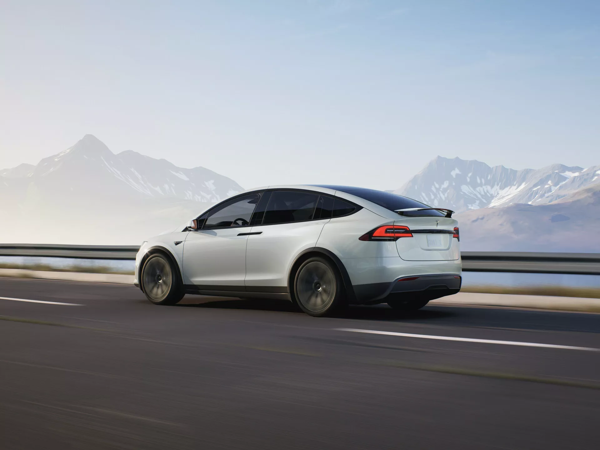 Dynamický pohľad na zadnú tretinu 2021 Tesla Model X (1. generácia-facelift) v pohybe, zvýrazňujúci jeho aerodynamický dizajn, elegantný biely lak, LED zadné svetlá a moderný dizajn kolies. Auto je zachytené pri pohybe zľava doprava proti rozmazanej ceste a zvodidlám, čo naznačuje rýchlosť, s horským horizontom v pozadí. Mäkké osvetlenie naznačuje skoré ráno alebo neskoré popoludnie, čo zdôrazňuje čisté elektrické línie a pokročilé funkcie vozidla v malebnom prostredí.