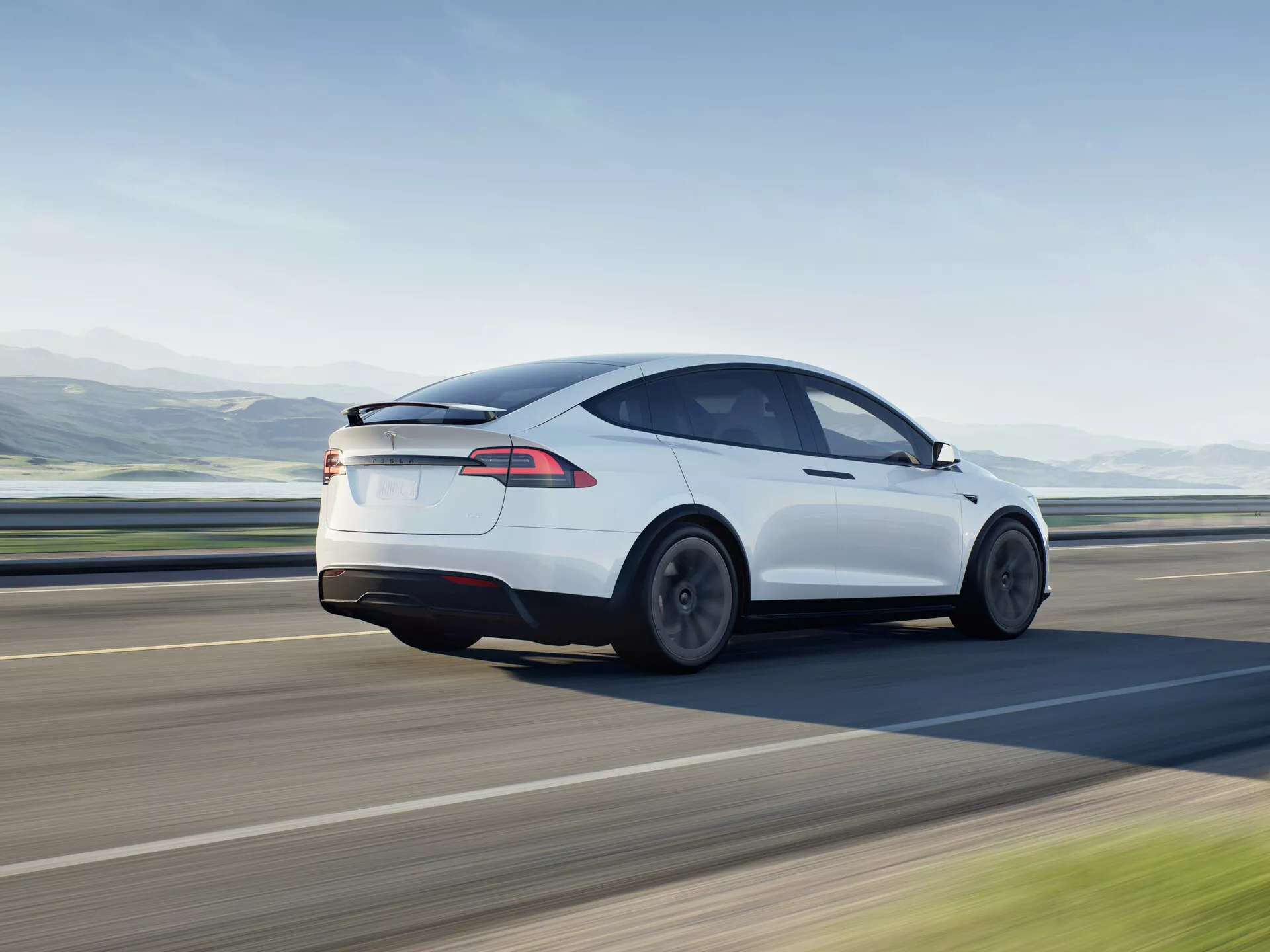 Dynamický obraz bieleho elektrického hatchback sedanu Tesla Model X 2021 v pohybe, zobrazený zo zadného trojštvrťového uhla. Fotografia zachytáva ľavý bok vozidla z mierne zvýšenej perspektívy, zdôrazňuje zadné kolesá, koncové svetlá a kontúru zadnej časti vozidla, s dôrazom na jeho aerodynamický dizajn so sklonenou strechou a hladkými krivkami. Dizajn naznačuje znížené vzdušné turbulencie a nízky koeficient odporu, podporený kolesami navrhnutými pre efektivitu aerodynamiky. Zadné okno splýva do čiernej sklenenej strechy, čo naznačuje panoramatické strešné okno, symbolizujúce modernú automobilovú technológiu. Elegantné LED koncové svetlá sú výraznou charakteristikou súčasnej osvetľovacej technológie. Absencia výfukového potrubia naznačuje plne elektrický charakter vozidla. Vhodné pre kategórie ako vozidlá, elektrické vozidlá, pohybová fotografia, automobilový dizajn, moderná technológia a luxusné vozidlá.