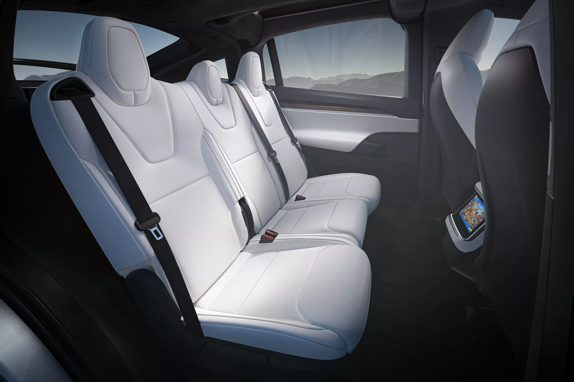Vnútorný záber na Tesla Model X 2021 (1. generácia-facelift), zobrazujúci zadný priestor pre cestujúcich zo zadných dverí na strane vodiča. Biele kožené sedadlá s opierkami hlavy a tvarovaným čalúnením pojmú troch pasažierov. Digitálny displej na zadnej strane sedadla predného spolujazdca naznačuje systém zadnej zábavy. Priestranná kabína, pravdepodobne s panoramatickým strešným oknom, využíva prirodzené svetlo na zvýraznenie kontrastných tmavých panelov dverí s integrovanými mriežkami reproduktorov.
