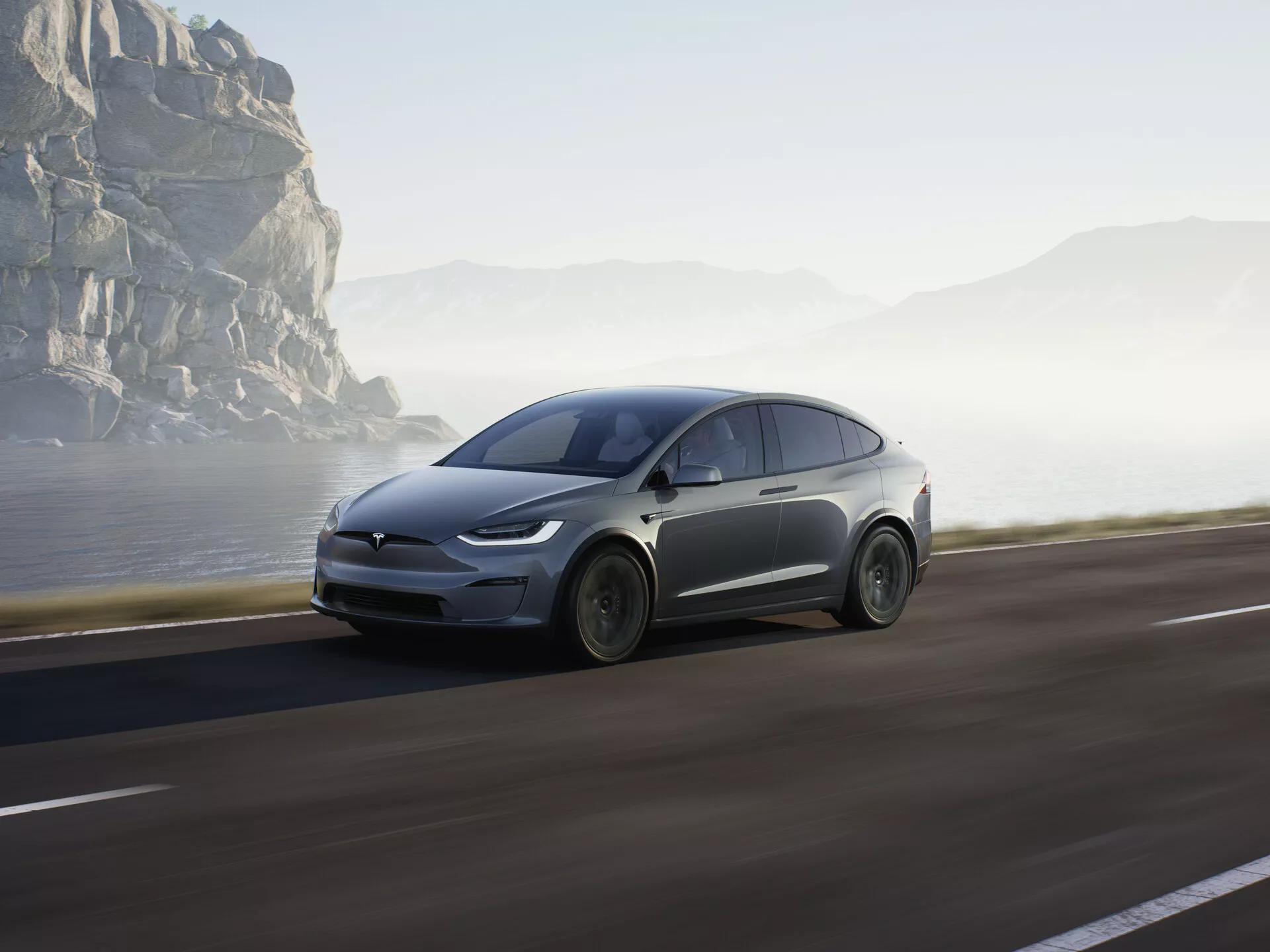 Obrázok ukazuje moderné elektrické SUV, konkrétne Tesla Model X 2021 (1. generácia facelift), idúce po pobrežnej ceste. Zobrazené je z nízkeho bočného profilu, ktorý zvýrazňuje prednú štvrtinu s dôrazom na predné koleso, kapotu, čelné sklo a časť dverí vodiča. Vozidlo zaberá podstatnú časť spodnej polovice rámu, čo zvýrazňuje jeho dominanciu. Auto je v pohybe s miernym rozmazaním kolies a pozadia, čo vytvára dojem rýchlosti a dynamiky. Mäkké slnečné svetlo naznačuje skoré ráno alebo neskoré popoludnie, čo pridáva scéne pokojnú náladu v malebnom a dobrodružnom pobrežnom prostredí. Lze predpokladať aerodynamické dizajnové prvky a pokročilé funkcie, ako sú LED svetlá, čo naznačuje pokročilú technológiu ako regeneratívne brzdenie a možno aj autonómne riadenie. Auto je umiestnené na pravej strane cesty a pohybuje sa vpred smerom k otvorenému cieľu, pričom pobrežné skaly a hmla v pozadí zvýrazňujú jeho futuristický vzhľad a prirodzené prostredie.