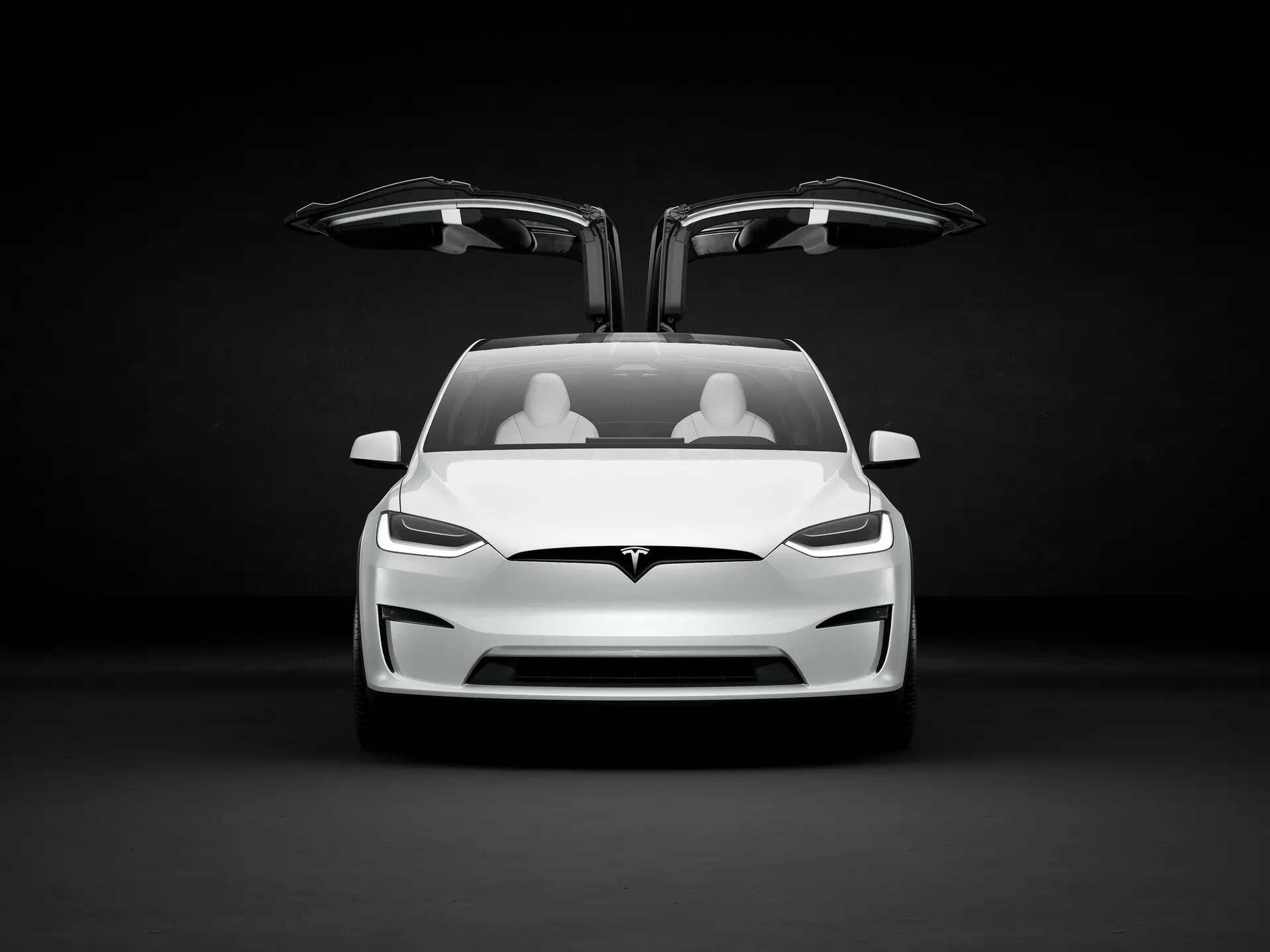 Obrázok zobrazuje elektrické vozidlo Tesla Model X 2021 (prvá generácia, facelift) v štúdiovom prostredí s tmavým pozadím, ktoré kontrastuje so svetlou farbou auta. Pohľad je mierne z nízkeho uhla a zvýrazňuje prednú časť auta, vrátane kapoty, nárazníka a oblasti mriežky, hoci vzhľadom na to, že ide o elektrické vozidlo, nemá tradičnú otvorenú mriežku. Viditeľný je aj dolný prívod vzduchu. Obe predné dvere sú otvorené, čím sa odhaľujú unikátne dvere typu falcon-wing, ktoré sa vyklápajú nahor, čo je typickým znakom luxusných modelov. Interiér ukazuje dve rady svetlých sedadiel, čo naznačuje minimalistický, ale luxusný dizajn. Ostré, hranaté LED svetlá dodávajú agresívny vzhľad a logo Tesla je výrazne umiestnené nad oblasťou prívodu vzduchu a pod predným kufrom - 'frunkom'. Auto naznačuje pokročilé technológie, ako sú autonómne riadenie, elektrický pohon, regeneračné brzdové systémy, konektivita a pokročilé bezpečnostné systémy, zamerané na publikum, ktoré ocení inovácie, výkon a ekologické povedomie.