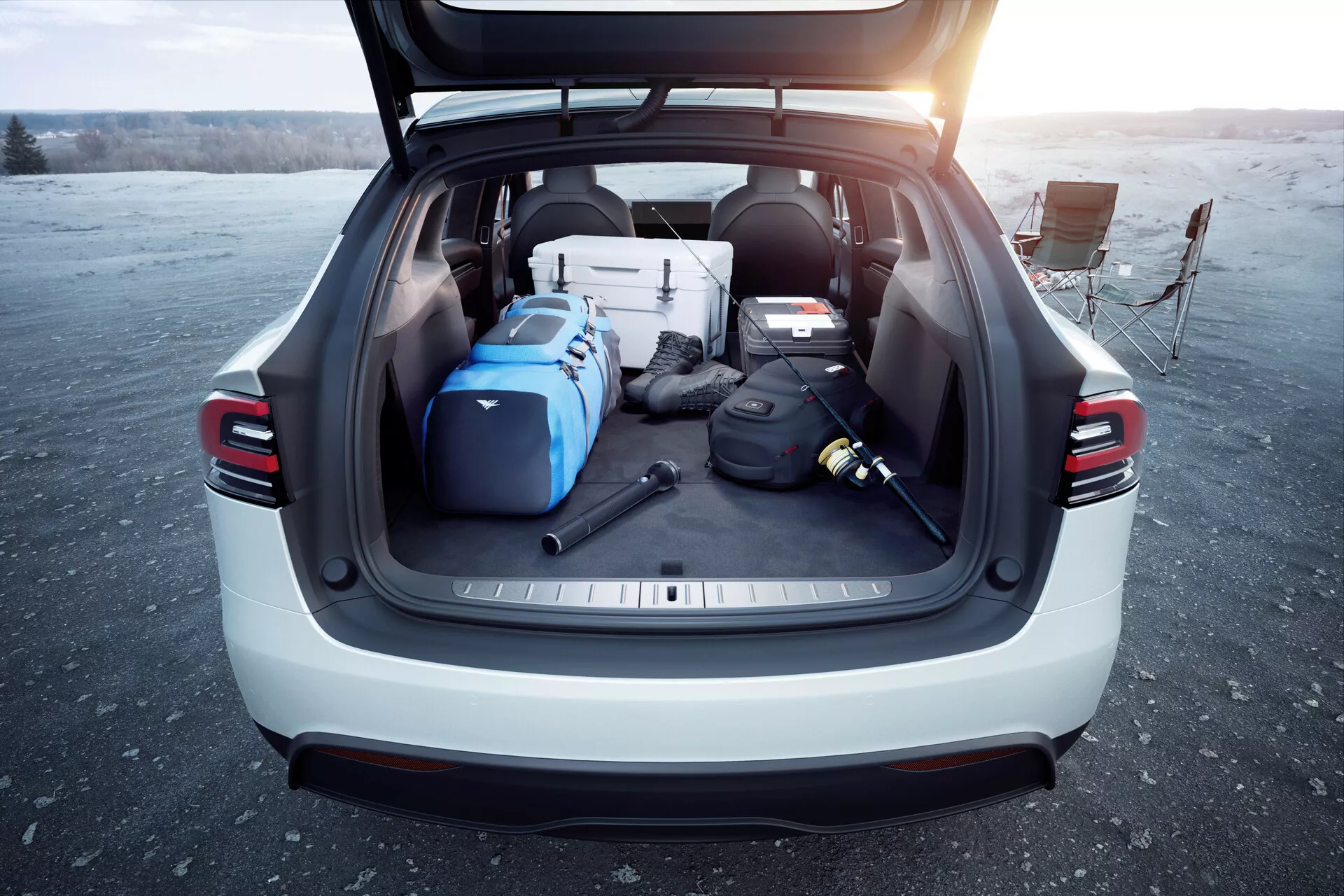 Zadný pohľad na SUV Tesla Model X 2021 s otvoreným kufrom, zobrazujúci priestrannú nákladnú časť plnú rôznych predmetov za denného svetla, pravdepodobne pri východe alebo západe slnka. Obrázok zdôrazňuje užitočnosť a moderné funkcie vozidla, ako sú automatické zadné dvere, zadné LED svetlá, svetlá farba exteriéru a kryt nákladu, fotené na otvorenom priestore.