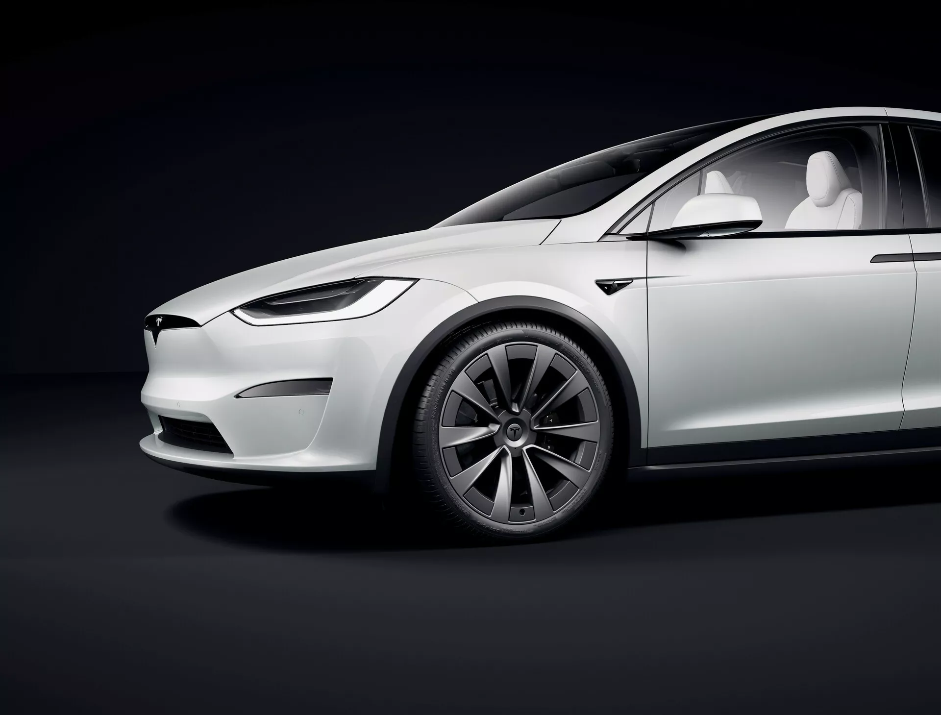 Štúdiový záber ukazujúci bočný profil vozidla Tesla Model X 2021 prvej generácie facelift z nízkeho uhla, zvýrazňujúci luxusné elektrické prvky, ako je elegantný dizajn, absencia výfukových rúr, minimalistický predok s LED svetlometmi, zasúvacie kľučky, viacramenné aero kolesá, veľké nízkoprofilové pneumatiky, lesklý biely lak s jemnými odrazmi, panoramatické strešné okno a kamery namiesto spätných zrkadiel. Krivky a línie vozidla sú zvýraznené štúdiovým osvetlením proti tmavému gradientnému pozadiu.