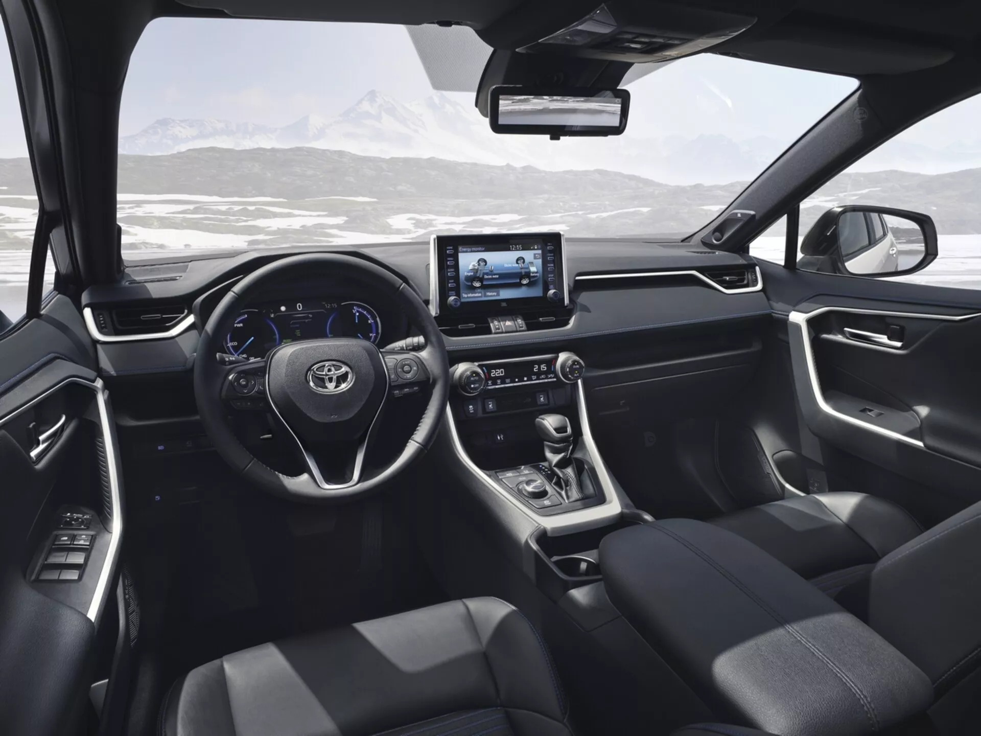 Interiér Toyota RAV4 2019 z pohľadu zo zadného sedadla, prezentujúci moderný dizajn a technologické prvky vrátane volantu s ovládacími tlačidlami, digitálneho displeja, dotykového infotainment systému, páky automatickej prevodovky, tmavo čalúnených sedadiel a ovládacích panelov na dverách s výhľadom na malebnú zasneženú horskú krajinu cez čelné sklo.