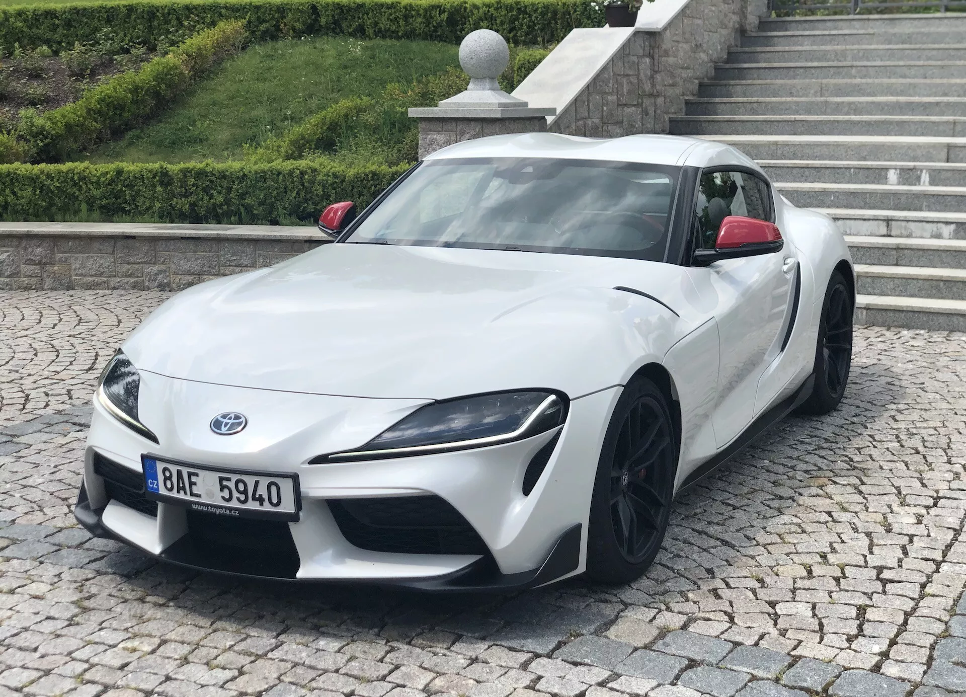 Obrázok zobrazuje biely Toyota Supra 2019 (toyota-gr-supra-2-0-pure-automatic) z predného trojštvrťového uhla. Jasne sú viditeľné nízky a široký postoj auta, ostré uhlové svetlá a čierne viacramenné hliníkové kolesá. Fotografia zdôrazňuje elegantný dizajn, aerodynamické kontúry a výkonnostnú estetiku. Športové auto je zaparkované na dlažbe s obrubníkom a schodiskom v pozadí, čo naznačuje mestské alebo obytné prostredie.