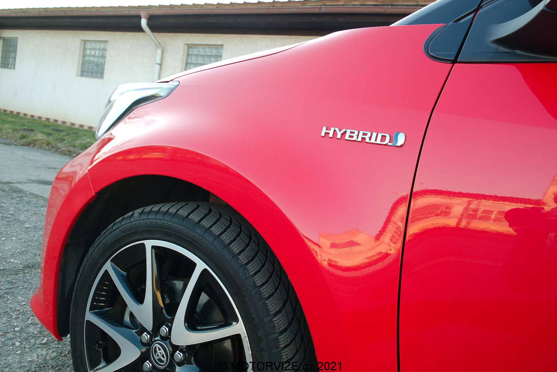 Detailný pohľad na červený Toyota Yaris 2020, zameraný na predný panel a viaclúčové zliatinové kolesá. Viditeľný je znak hybridu, zdôrazňujúci ekologickú technológiu vozidla. Lesklý lak odráža slnečné svetlo, zdôrazňujúc vysokú kvalitu dokončenia a moderný, aerodynamický dizajn auta.