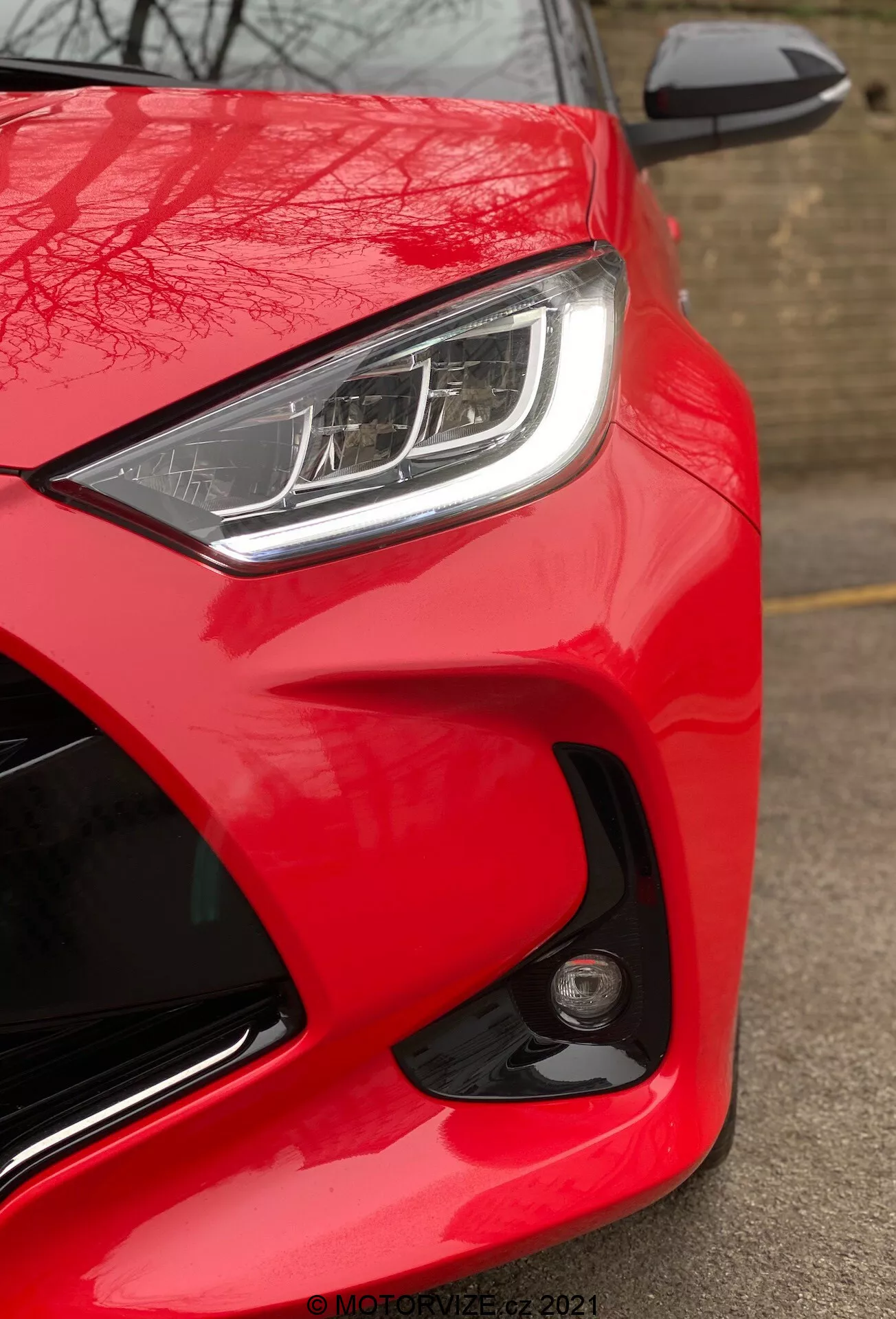 Detailný pohľad na Toyota Yaris 2020, zameraný na prednú časť zo strany vodiča z nízkeho uhla. Obrázok zvýrazňuje jasnú, lesklú červenú farbu automobilu, elegantné LED alebo HID svetlá s výraznými dennými svetlami a čiastočne viditeľné zrkadlo vo farbe karosérie s integrovaným ukazovateľom smeru. Aerodynamický dizajn je zdôraznený tvarovanými líniami a krivkami okolo svetiel a kapoty. Okrúhle hmlové svetlo alebo senzor na prednom nárazníku naznačuje pokročilé bezpečnostné funkcie. Absencia rozmazania pohybu naznačuje, že auto je statické, a v pravom dolnom rohu je viditeľný vodoznak 'MOTORZETZ. 2021'.