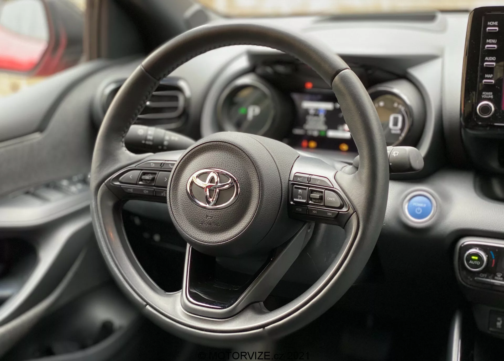 Pohlad do interiéru Toyoty Yaris 2020 zameraný na volant a palubnú dosku z perspektívy vodičovho sedadla. Volant má značku Toyota a viditeľné ovládacie prvky pre infotainment a asistenčné systémy vodiča. Za ním sú viditeľné časti digitálneho prístrojového panela a stredovej konzoly s ovládacími prvkami klimatizácie a výduchmi.