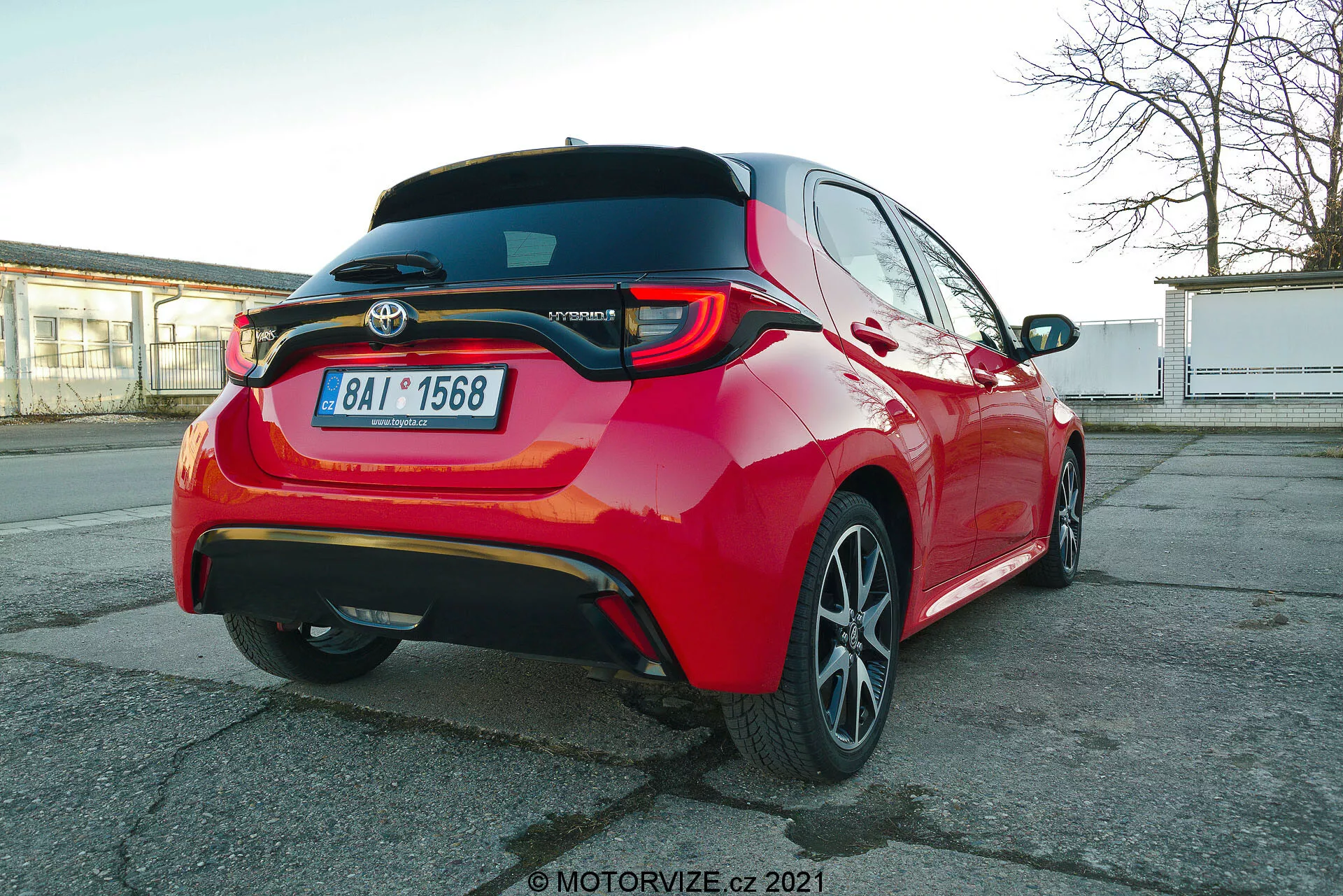 Živo červené osobné auto Toyota Yaris 2020, fotografované zo zadného trojštvrťového uhla, zvýrazňujúce LED zadné svetlá, zadný spojler, emblém modelu Hybrid, viacramenné zliatinové ráfiky a viditeľné asistenčné funkcie vodiča, ako sú parkovacie senzory a zadná kamera, zachytené z perspektívy mierne pod úrovňou výšky auta.