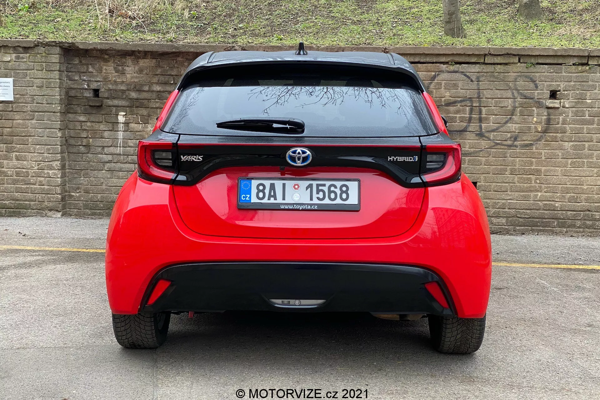 Zadný pohľad na červený Toyota Yaris Hybrid 2020, vycentrovaný a nasnímaný priamo. Obrázok odhaľuje celú šírku vozidla, zobrazuje logo Toyota, modelové názvy 'Yaris' a 'Hybrid', spojler, anténu namontovanú na streche, vysoko umiestnené brzdové svetlo a výrazné zadné svetlá. Viditeľné je aj zadné okno so stieračom a spodná časť nárazníka, ktorá pravdepodobne obsahuje senzory alebo odrazky. Na pozadí je tehlová stena alebo plot a nejaká vegetácia, čo naznačuje nižšie zaparkované auto alebo nižší uhol kamery.