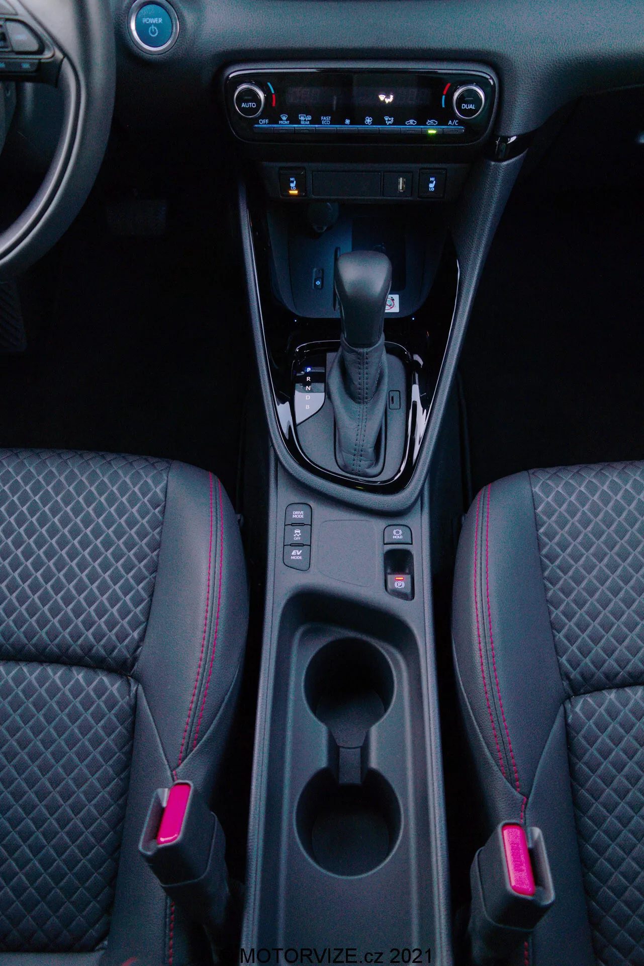 Vertikálny záber interiéru Toyota Yaris 2020 zameraný na stredovú konzolu, ktorá obsahuje radiacu páku v polohe 'P', tlačidlo elektronickej parkovacej brzdy, dve osvetlené držiaky na poháre, úložný priestor, digitálnu klimatizáciu, dotykový multimediálny systém, USB port, vetracie otvory a dvojfarebné predné sedadlá so vzorovaným prešívaním a červeným akcentom na prešívaní.
