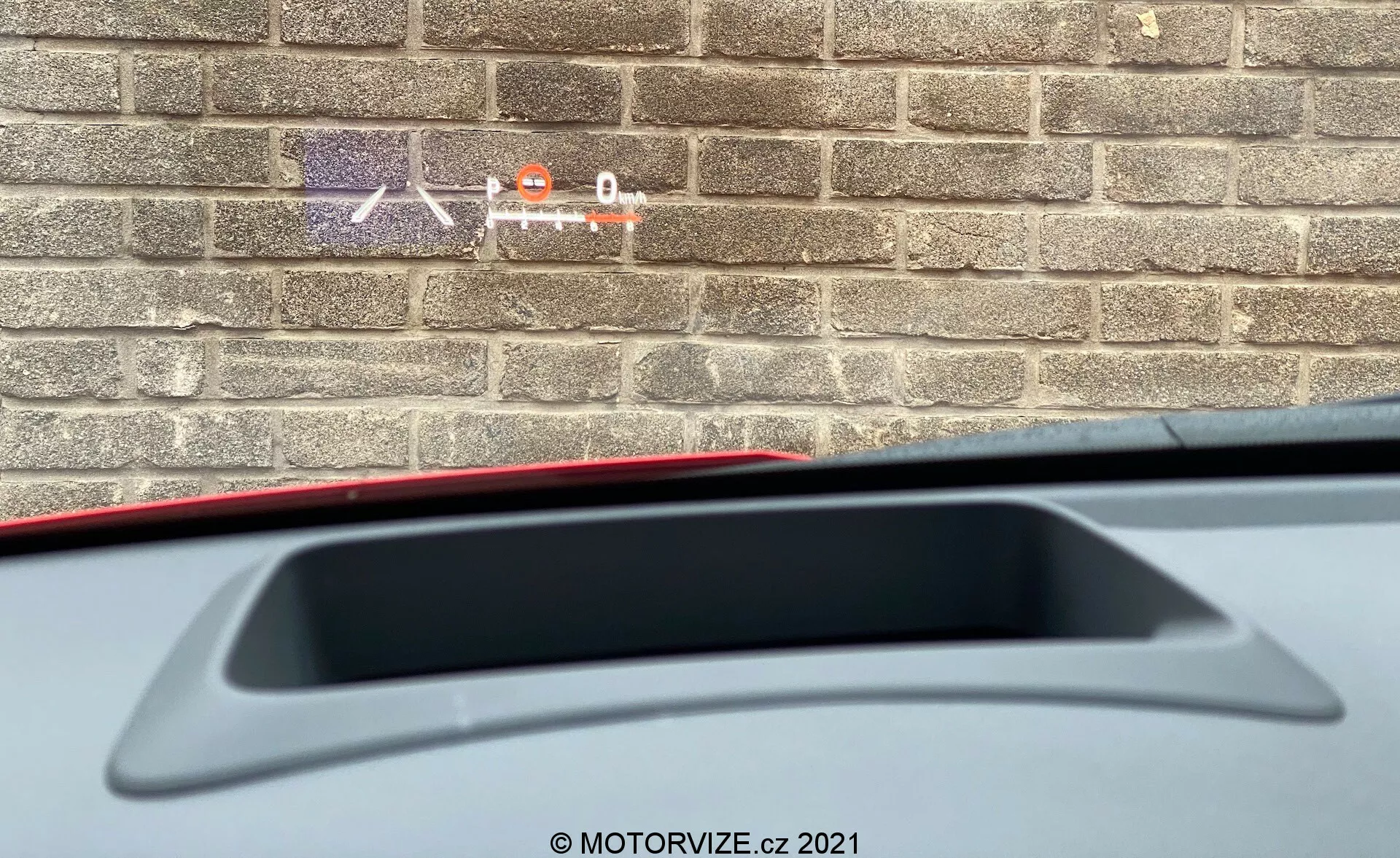 Detailný pohľad zvnútra Toyota Yaris 2020 ukazujúci čiastočnú perspektívu z vodičovho sedadla smerom dozadu. Zameranie je na zadnú odkladaciu poličku s vysoko umiestneným brzdovým svetlom a integrovaným zadným spojlerom. Obrázok tiež zobrazuje časť materiálu interiéru, pričom tehličková stena vonku naznačuje, že auto je zaparkované blízko budovy.