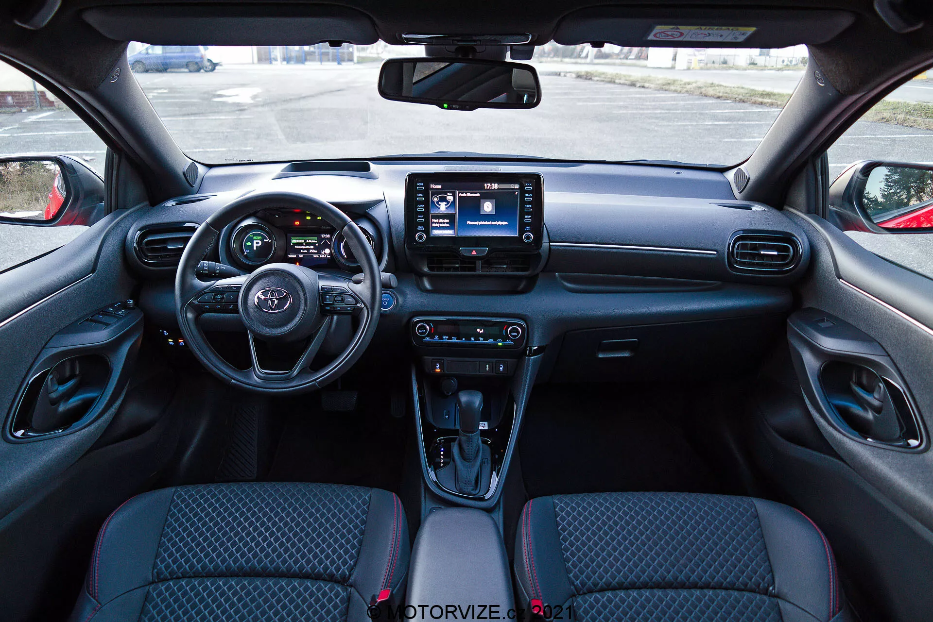 Pohľad na interiér vozidla Toyota Yaris 2020 z pohľadu sedadla vodiča, zobrazujúci modernú prístrojovú dosku s volantom, ktorý má integrované ovládacie prvky, čiastočne viditeľný prístrojový panel s digitálnymi obrazovkami a centrálny infotainment systém s dotykovým displejom. Prístrojová doska obsahuje ventilačné otvory a ovládacie prvky klimatizácie, zatiaľ čo stredová konzola obsahuje ovládacie prvky režimov jazdy, automatickú rýchlostnú páku s koženým poťahom, držiaky na poháre a úložný priestor. Sedadlá s látkovým čalúnením majú vzorovaný dizajn a vnútorné panely dverí obsahujú ovládacie prvky pre elektrické okná a nastavenie zrkadiel.