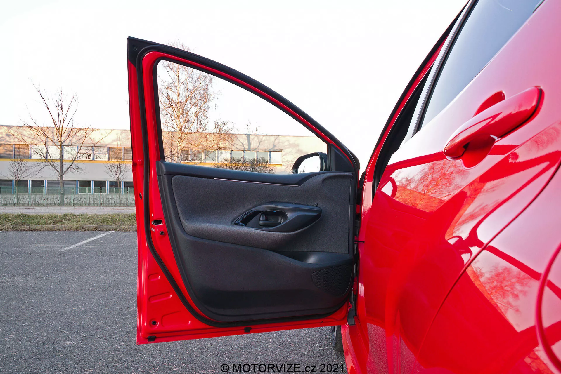 Detailný pohľad na červený Toyota Yaris 2020 zo strany vodiča s otvorenými prednými dverami, zachytený z mierneho nízkeho uhla. Kamera je umiestnená vonku, blízko dverí, smerujúc dovnútra, ukazujúc jasný červený lak odrážajúci svetlo a čierny vnútorný dekor s minimalistickým dizajnom. Dvere majú tlačidlo na ovládanie elektrických okien vo vnútri rukoväte a vrecko na uloženie pod ním.