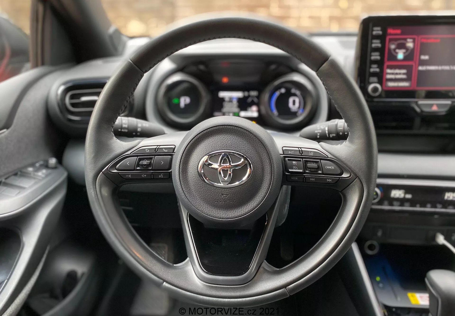 Detailný pohľad na interiér Toyota Yaris 2020 zo sedadla vodiča, s trojramenným volantom s emblémom Toyoty a ovládacími tlačidlami pre audio, hands-free volanie a tempomat. Digitálny prístrojový panel zobrazujúci rýchlosť a základné informácie o jazde je čiastočne viditeľný. Tiež viditeľná stredová konzola s infotainment dotykovou obrazovkou zobrazujúcou možnosti ponuky.