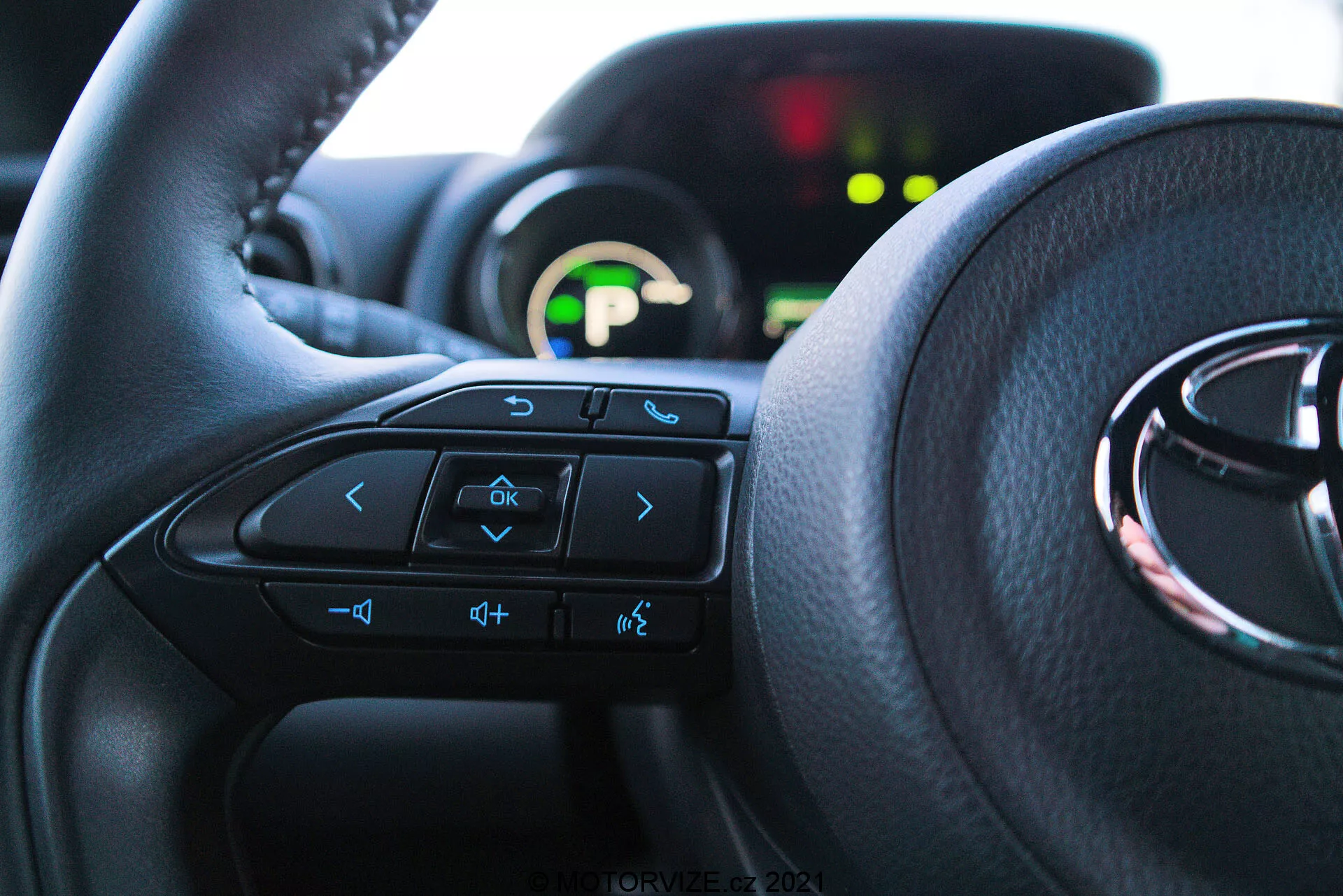 Blízky interiérový pohľad na Toyota Yaris 2020 z perspektívy sedadla vodiča, zvýrazňujúci volant s multifunkčnými ovládačmi a prístrojovým panelom v pozadí. Volant dominuje dolným dvom tretinám snímky, viditeľné tlačidlá zahŕňajú šípky, hlasové ovládanie, ovládanie hlasitosti a zábavného systému. Čiastočne viditeľný prístrojový panel je osvetlený zeleným digitálnym displejom a je rozostrený kvôli nízkej hĺbke ostrosti. Interiér je osvetlený prirodzeným svetlom, čo zdôrazňuje pohodlie a užívateľsky prívetivé rozhranie.