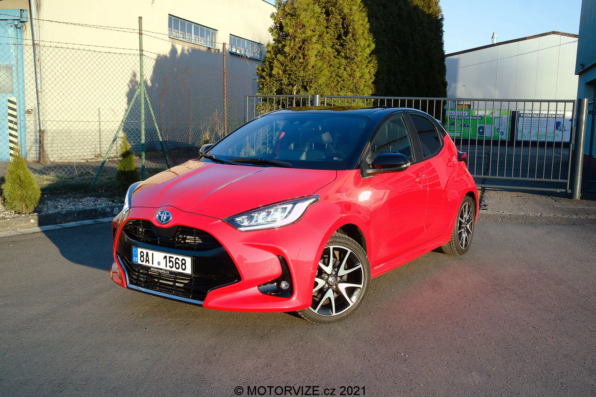 Obrázok jasne červeného hatchbacku Toyota Yaris z roku 2020 zaparkovaného pod uhlom, ktorý ukazuje ako predný, tak bočný profil. Dizajn svetiel je ostrý a uhlový, mriežka má vzor včelieho plastu s centrálnym emblémom a predný nárazník má viditeľné kontúry s hmlovými svetlami. Bočný profil odhaľuje dvojfarebné zliatinové kolesá, farebne zladené kľučky dverí a zrkadlá s ostrými líniami a kontúrami pozdĺž boku. Lesklý lak naznačuje nový alebo dobre udržiavaný stav. Pozadie zahŕňa jasnú oblohu, priemyselné štruktúry a mäkké tiene, čo naznačuje vonkajšie prostredie, pravdepodobne parkovisko v blízkosti komerčného alebo priemyselného zariadenia.