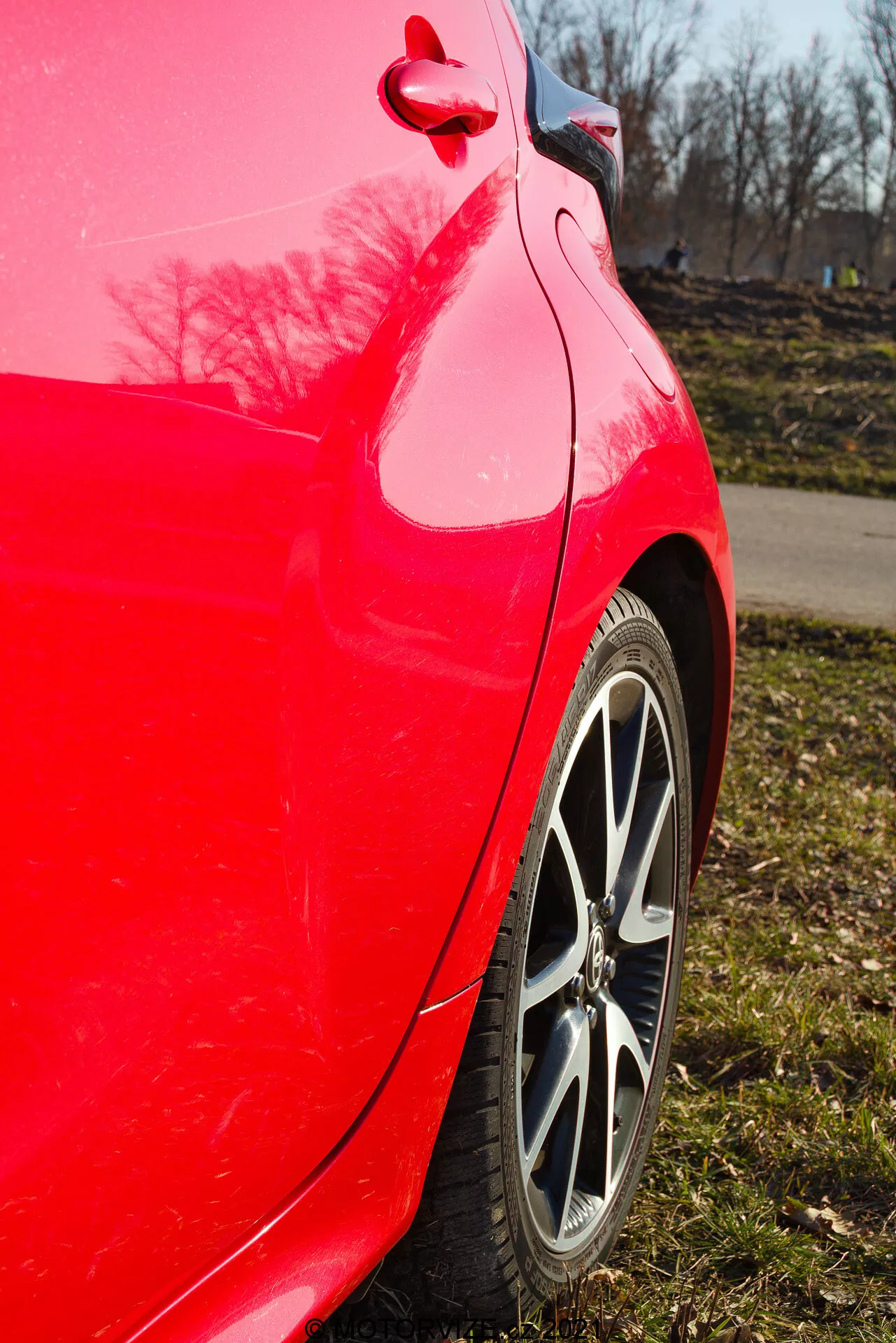Zblízka bočný pohľad na zadnú štvrtinu panelu červeného auta Toyota Yaris 2020, zachytený v mierne vzostupnom uhle, zvýrazňujúc jasný červený lak, viacpaprskový zliatinový ráfik, vzor dezénu pneumatík, čistú zakrivenosť zadného štvrťpanelu a nárazníka, lesklý odraz farby so stromami a jasným modrým nebom, a viditeľný viečko palivovej nádrže a detailné obloženie.