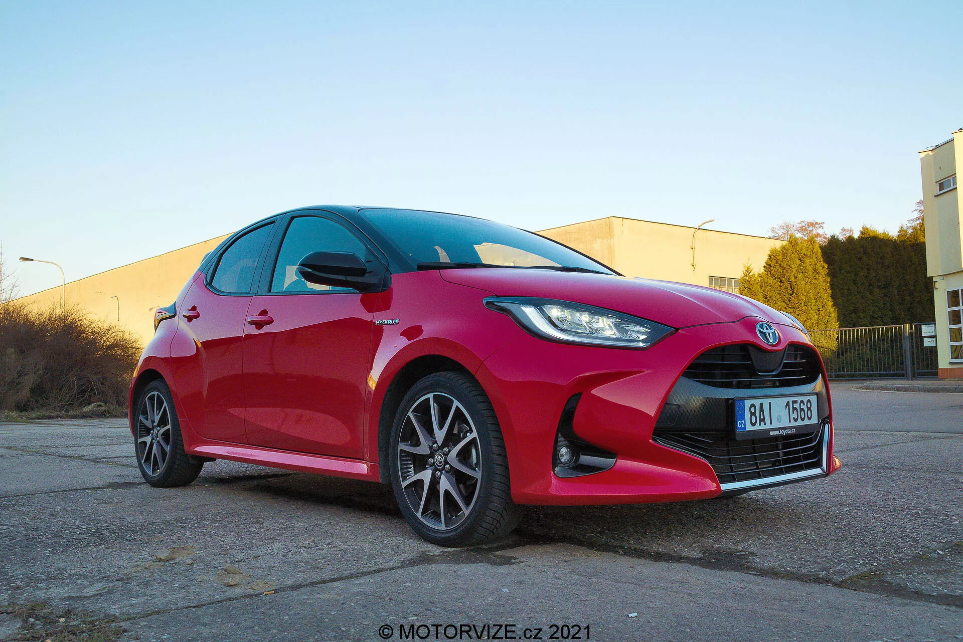 Nízkoúhlový pohľad spredu na tri štvrtiny zaostrený na živý červený Toyota Yaris hatchback 2020, ktorý zobrazuje ostré a uhlové LED alebo HID svetlomety, výraznú mriežku s centrálnym emblémom, viaclúčové zliatinové kolesá, integrované smerovky v spätných zrkadlách, čisté odrazové povrchy a pravdepodobne výkonný brzdový systém. Zachytené na teplo osvetlenom priemyselnom parkovisku s vodoznakom motorvize.sk 2021.