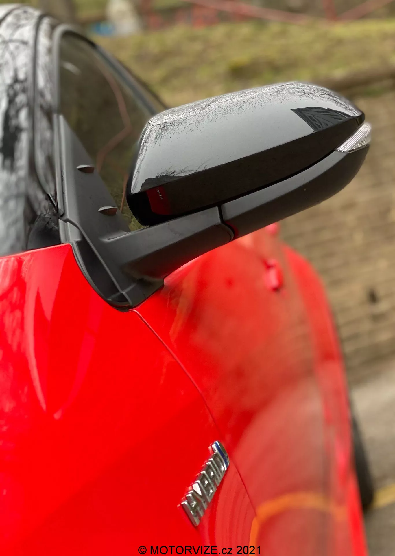 Detailný záber na vysunuté bočné zrkadlo na Toyote Yaris 2020 s elegantným, aerodynamickým čiernym krytom kontrastujúcim s lesklou červenou karosériou auta. Zrkadlo zdôrazňuje technologické prvky ako možné elektrické nastavenie, vyhrievanie a integrované smerovky, aj keď konkrétne funkcie nie sú potvrdené. Kvapky vody naznačujú nedávny dážď alebo umytie auta, zatiaľ čo rozmazané pozadie a plytká hĺbka ostrosti udržujú pozornosť na zrkadle. Čiastočne je vidieť odznak na prednom blatníku, ale ten je rozmazaný.