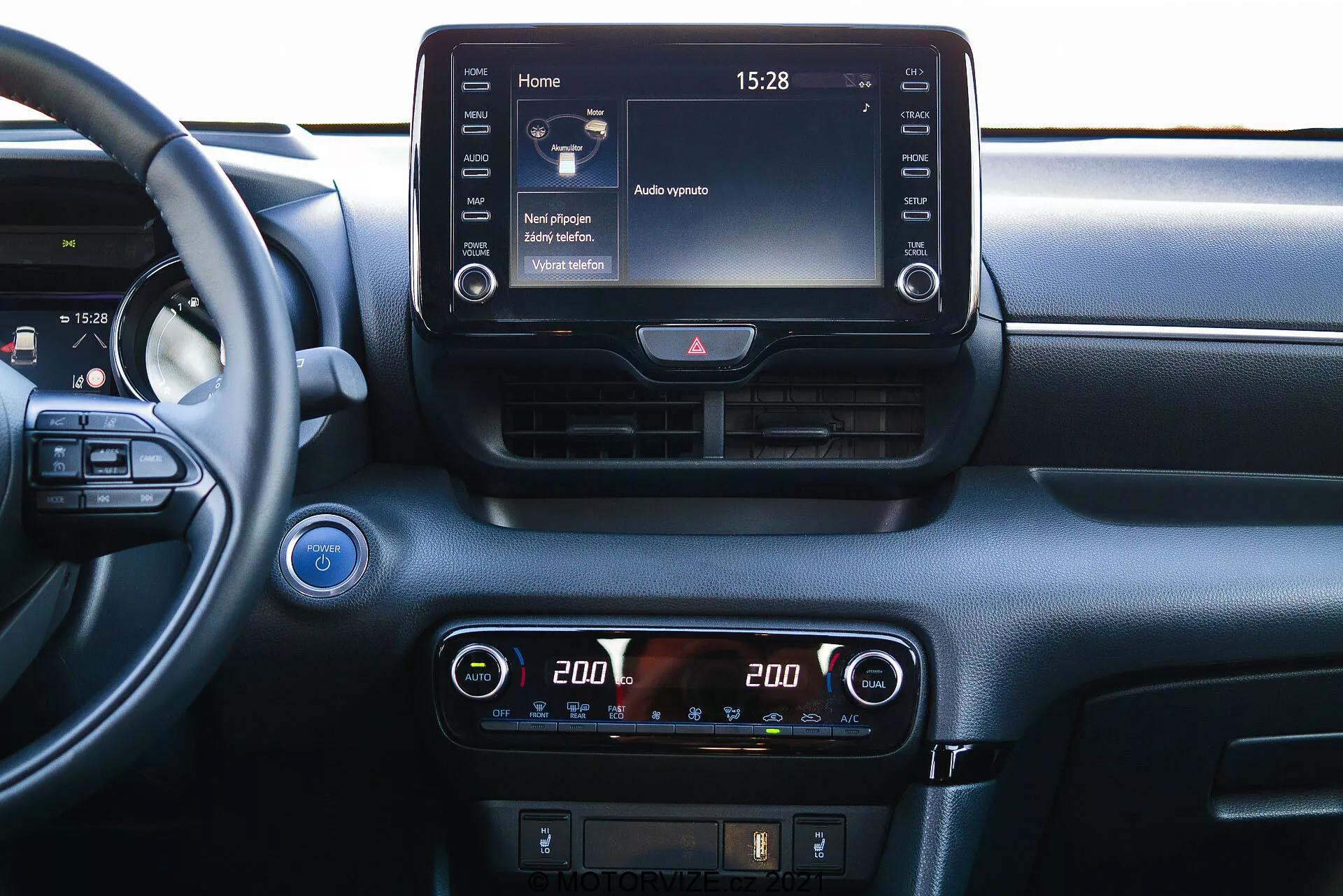 Detailný pohľad na centrálnu palubnú dosku modelu Toyota Yaris 2020 z perspektívy interiéru. Volant je čiastočne viditeľný vľavo, čo naznačuje uhol pohľadu z miesta spolujazdca alebo centra vozidla. Palubná doska obsahuje výrazný dotykový displej infotainmentu na vrchu stredovej konzoly, zobrazujúci domovskú obrazovku s rôznymi ikonami a čas '15:28'. Pod ním je digitálny displej klimatizácie s nastaveniami dvojzónovej teploty, každá strana nastavená na '20.0' stupňov Celzia. Pod ovládačmi klimatizácie sú viditeľné ďalšie tlačidlá na odmrazovanie a možno aj ovládanie asistenčných systémov vodiča. Nad displejom infotainmentu sú dve horizontálne vetracie otvory s tlačidlom výstražných svetiel medzi nimi. Na ľavom okraji je čiastočne viditeľné tlačidlo štart/stop, čo naznačuje technológiu bezkľúčového zapaľovania. V ľavom hornom rohu sa nachádza mriežka reproduktora. Materiál palubnej dosky obsahuje hladké aj textúrované prvky, čo naznačuje dôraz na pohodlie a technológiu.
