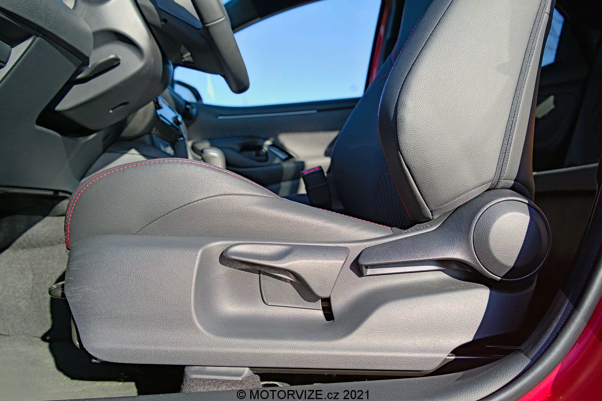 Detailný vnútorný pohľad na Toyota Yaris 2020 zameraný na dvere a sedadlo vodiča, zdôrazňujúci dvojfarebné čalúnenie, dekoratívne šitie, tvarovaný dizajn pre ergonomickú podporu, manuálne nastaviteľné bočné opierky a výraznú páku na manuálnu reguláciu sedadla. Rám dverí je čiastočne viditeľný, čo naznačuje, že dvere sú otvorené.