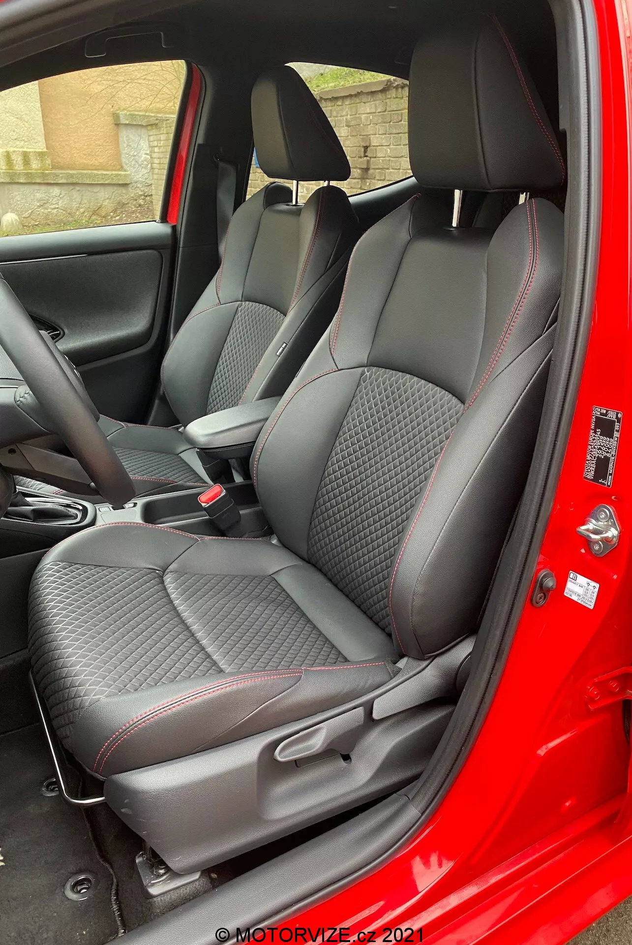 Interiér modelu Toyota Yaris 2020 na pohľad z miesta spolujazdca s otvorenými dverami, zvýrazňujúci prémiové textilné sedadlá so vzorom diamantu a červeným akcentom, nastaviteľné opierky hlavy, ovládací panel stredovej konzoly a panel dverí s textúrou z karbonových vlákien a elektrické ovládanie okien.