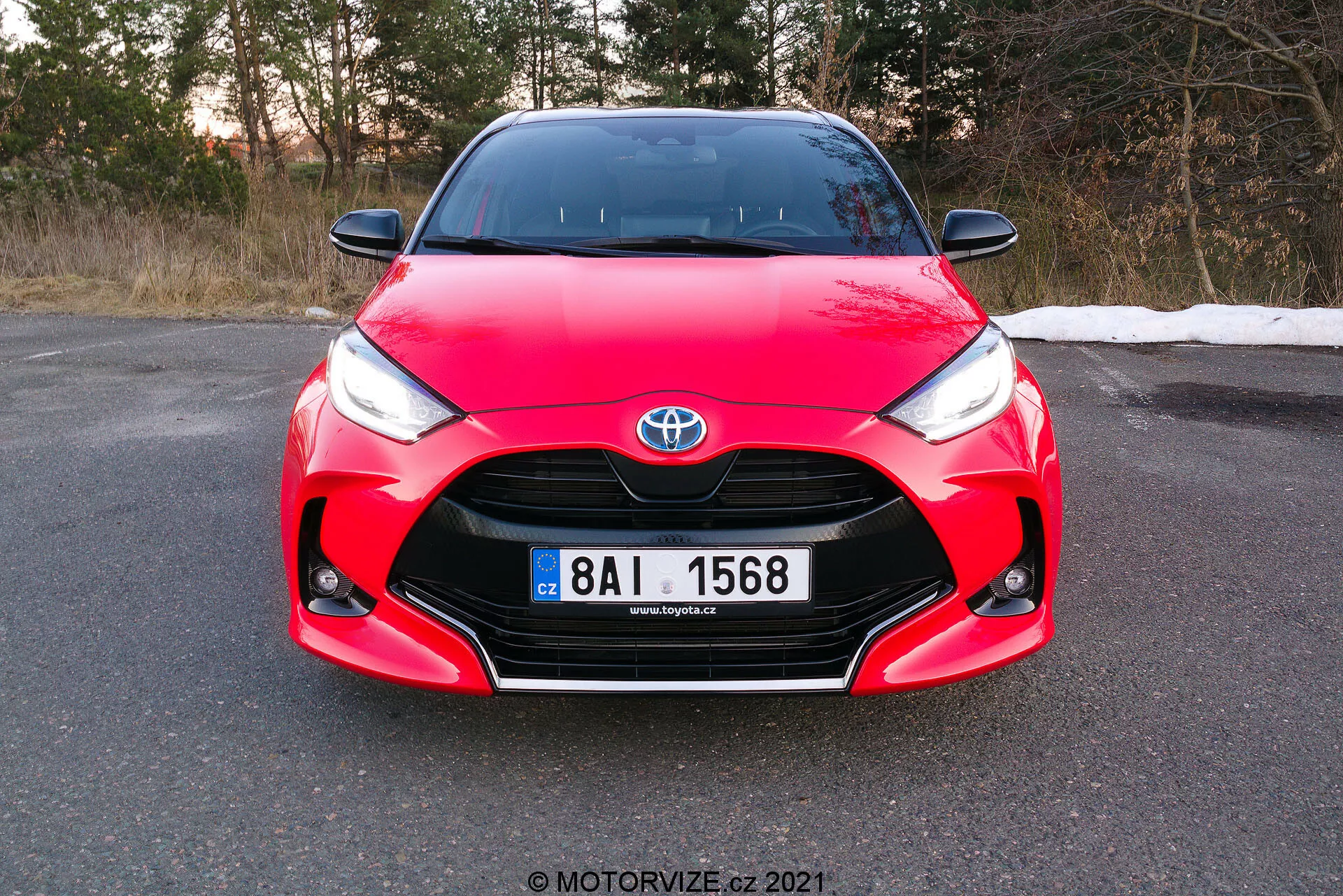 Obrázok zobrazuje predný pohľad na červený Toyota Yaris 2020, urobený pod miernym uhlom, zameraný na prednú masku, svetlomety a kapotu s úplne viditeľnou registračnou značkou. Automobil má charakteristické dizajnové prvky vrátane znaku Toyota uprostred mriežky, ostré hranaté svetlomety zužujúce sa k mriežke a zložité krivky na kapote, ktoré ukazujú jeho aerodynamický a športový dizajn. Fotografia urobená počas dňa pred súmrakom naznačuje, že vozidlo stojí na rovinatom povrchu v prostredí, ktoré by mohlo byť neskoro na jeseň alebo skoro na jar.