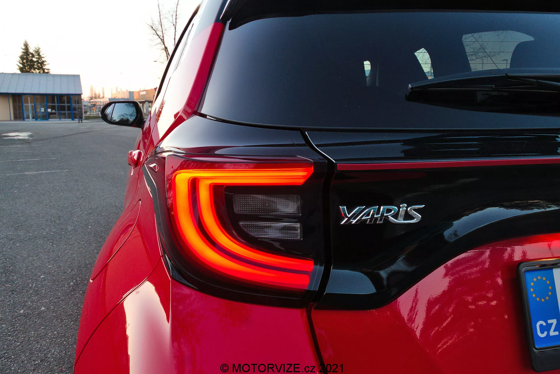 Detailný záber na zadnú časť červeného modelu Toyota Yaris z roku 2020, zobrazujúci ľavé zadné LED svetlo s výrazným tvarom C, názov modelu 'YARIS' na zadných dverách a poznávaciu značku Európskej únie z Českej republiky. Snímka v šikmom uhle pri súmraku zvýrazňuje obrysy a lesk auta, s rozostreným pozadím, ktoré sústreďuje pozornosť na dizajnové detaily.