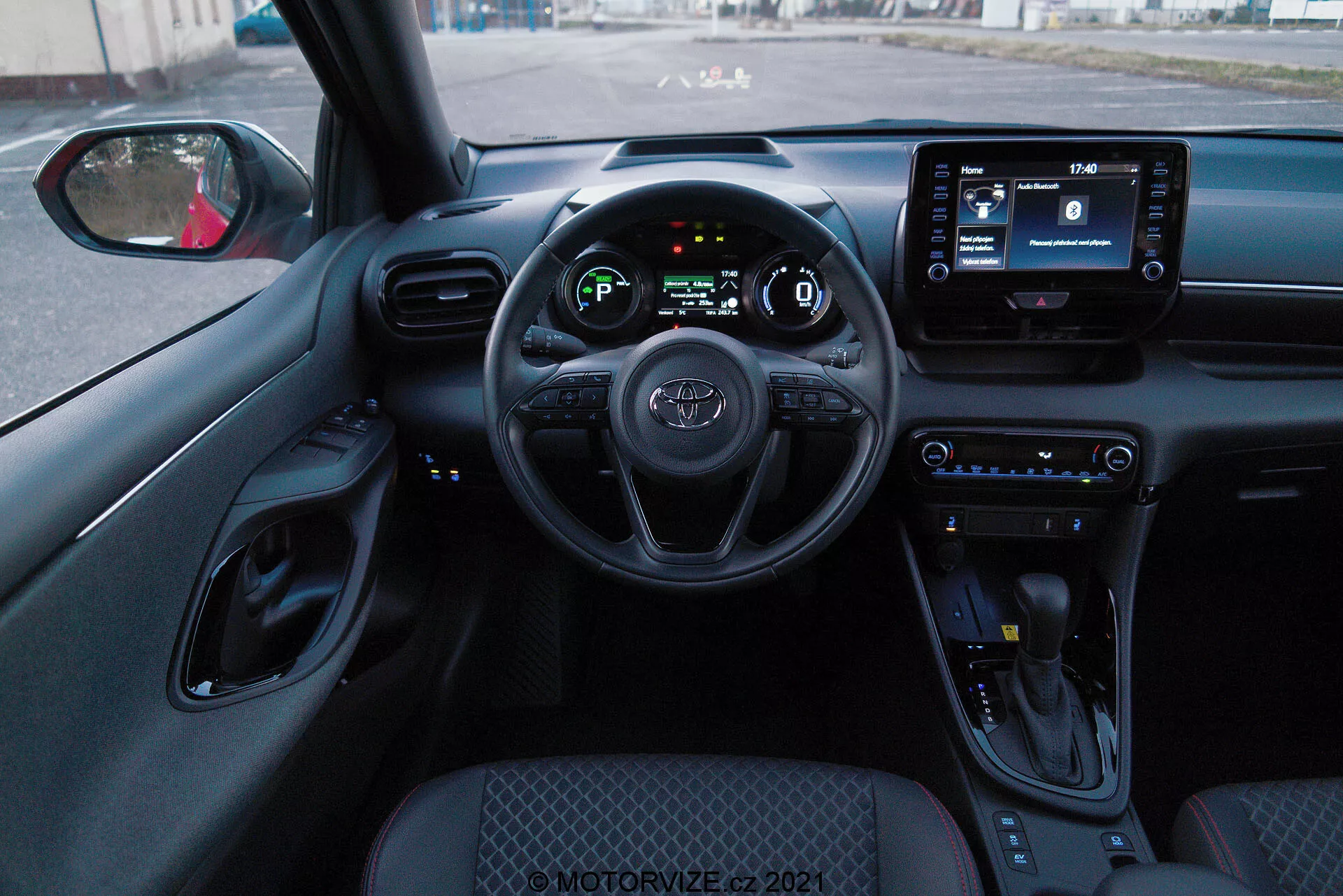 Interiérová perspektíva zo sedadla vodiča Toyoty Yaris 2020, ukazujúca volant s namontovanými ovládacími prvkami, digitálnu prístrojovú dosku a infotainmentový displej. Interiér má ovládacie tlačidlá klímy, radiacu páku a prevažne čierne farebné schéma s textúrovanými povrchmi a chrómovými akcentmi.