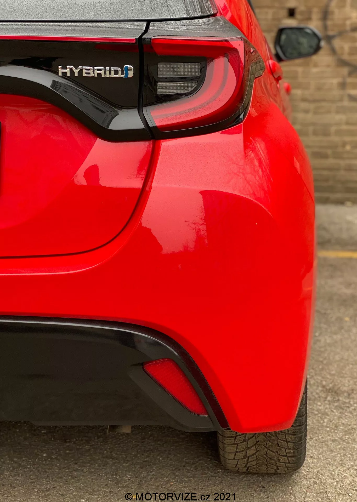 Detailný pohľad na zadnú časť červeného hybridného vozidla Toyota Yaris 2020 z uhla, zameraný na pravý zadný štvrtinový panel. Zachytáva prvky ako označenie 'HYBRID', moderné LED zadné svetlá, jemný aerodynamický záhyb na zadnom nárazníku a viditeľný parkovací senzor. Reflexný, lesklý lak a dobre udržiavaný dezén pneumatiky naznačujú, že vozidlo je nové alebo dobre udržované.