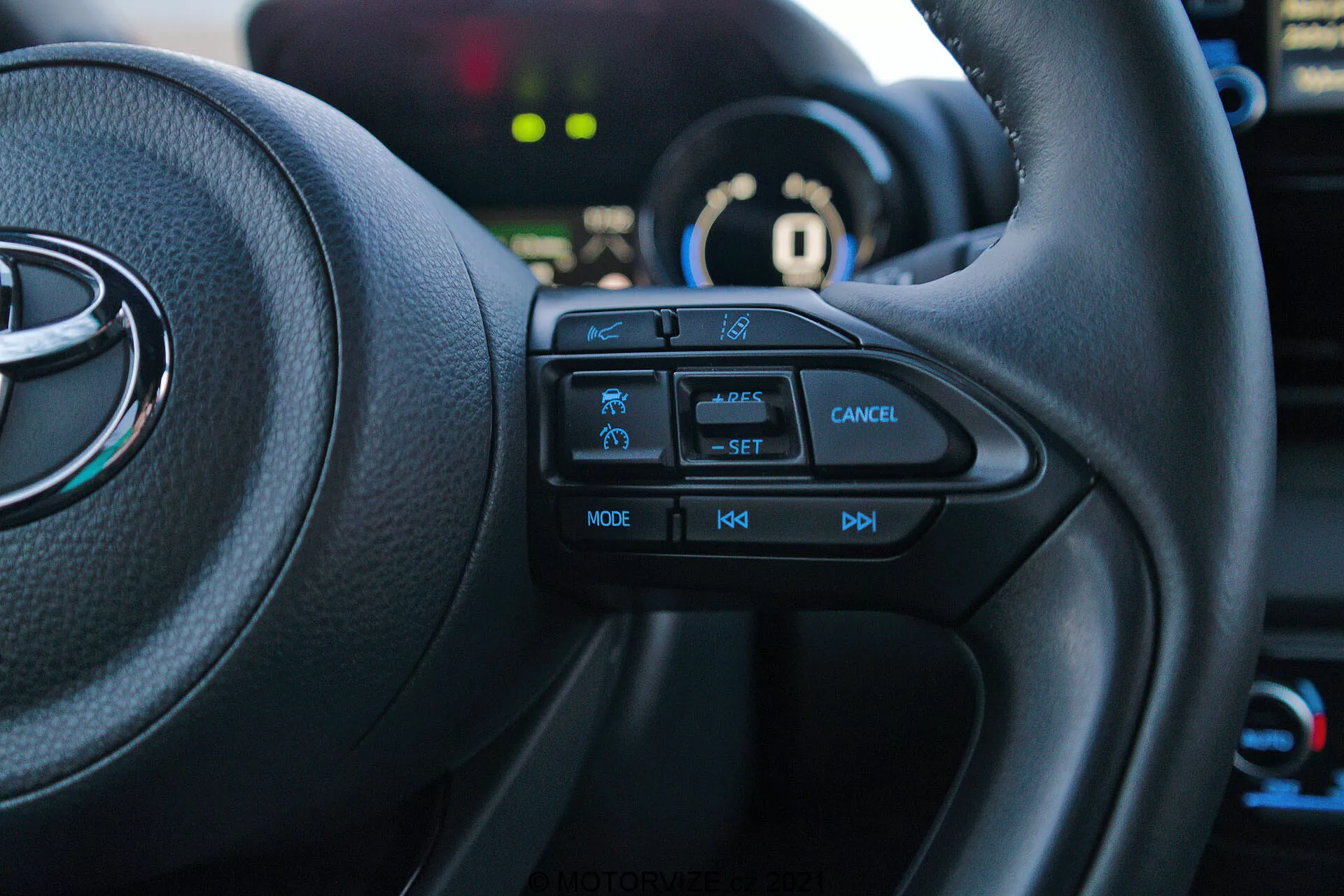 Interiérová snímka Toyota Yaris 2020 z pohľadu vodiča, zameraná na ľavú stranu multifunkčného volantu. Hmatateľné tlačidlá, vrátane ikony bezpečnostnej funkcie, tempomatu s označením 'SET', tlačidla zrušenia, 'MODE' pre multimédiá a ovládania hlasitosti '+', '-', sú jasne viditeľné. Vozidlo je v režime 'P' parkovanie, pričom prístrojový panel a pozadie sú mierne rozmazané.