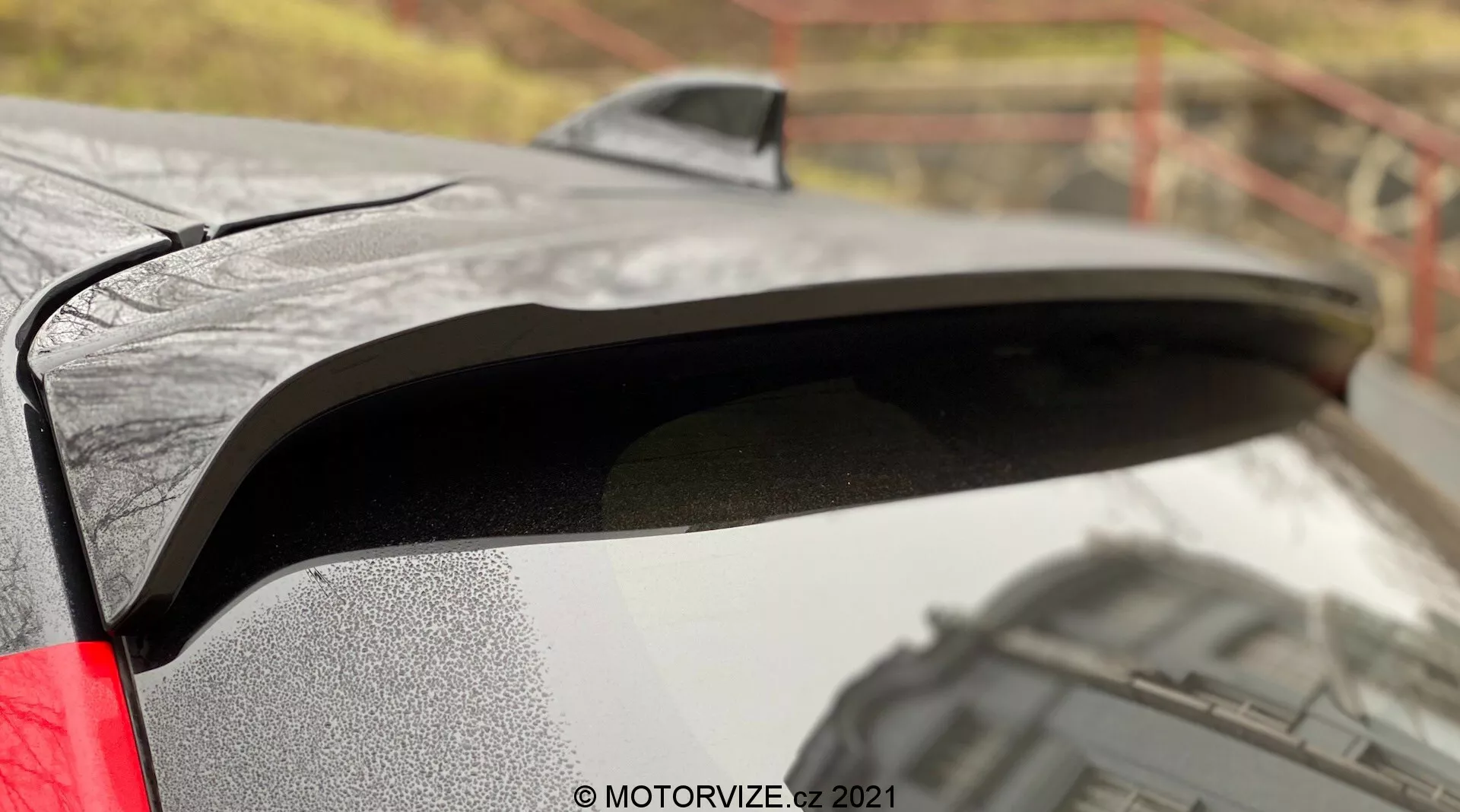 Detailný pohľad na zadný spojler Toyoty Yaris 2020 s čiernym náterom, ktorý kontrastuje s kovovým telom. Obrázok zachytáva kvapky vody na spojleri, čo naznačuje mokré podmienky, a bočný profil odhaľujúci mierne zdvihnutý dizajn pre lepšiu aerodynamiku. Rozmazané vonkajšie pozadie pridáva kontext k prémiovému dokončeniu auta a viditeľné zadné svetlo LED naznačuje moderné funkcie.