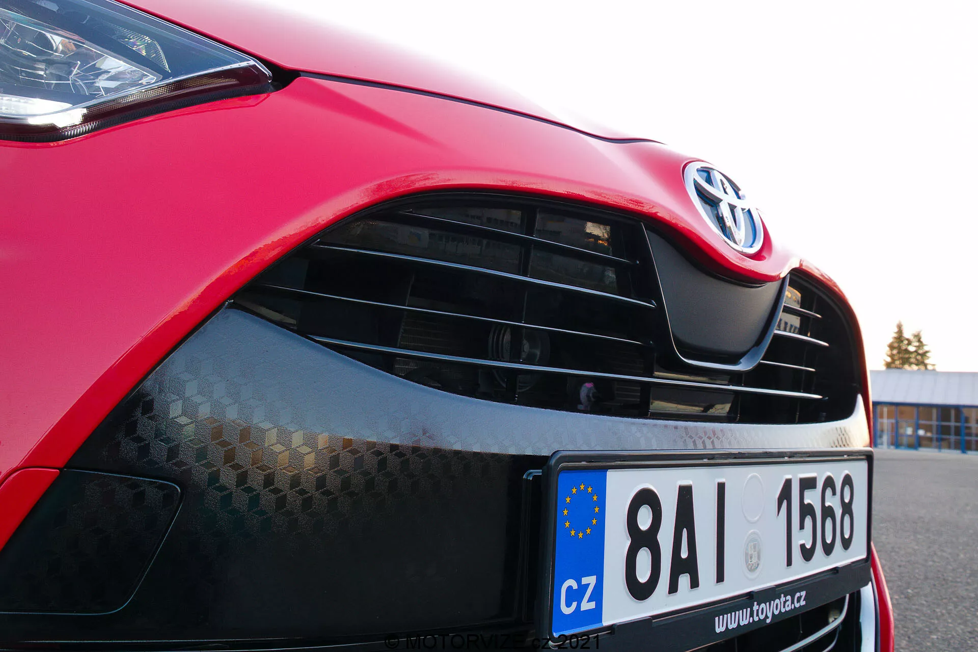 Detailný záber na červený Toyota Yaris 2020 zameraný na prednú ľavú stranu, predný kapotu, moderný svetlomet s možnou LED alebo HID technológiou, mriežku s emblémom Toyota a štruktúrou voštiny, lesklý červený lak a čiastočne viditeľný predný nárazník s dolným prívodom vzduchu. Európska registračná značka naznačuje registráciu v Českej republike.