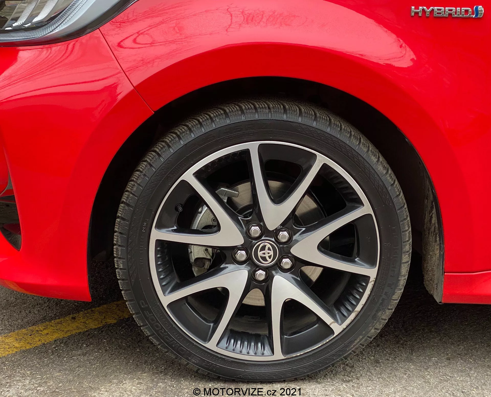 Záber na predné koleso a časť bočnej strany červeného hybridného auta Toyota Yaris 2020, fotografovaný z úrovne zeme. Viaccípe koleso z hliníkovej zliatiny má strieborné a čierne prevedenie. Vzor pneumatiky je jasne viditeľný, bez známok opotrebenia. Na boku pneumatiky sú vytlačené špecifikácie a nad kolesom na bočnej strane auta je viditeľný znak 'HYBRID'. Jednotné denné svetlo zvýrazňuje viditeľnosť dizajnu kolesa a hybridného označenia.