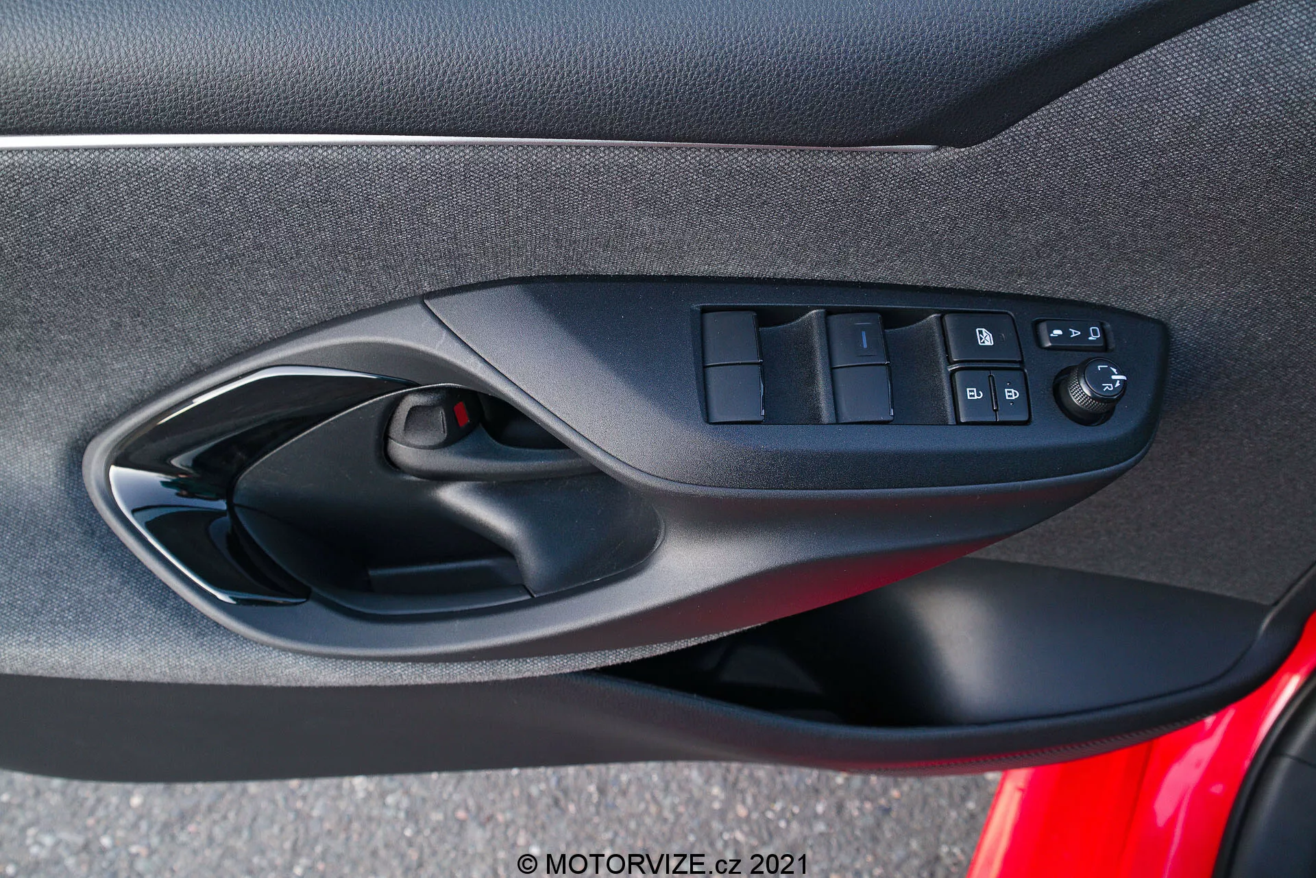 Interiérový dverný panel Toyoty Yaris 2020, zameriavajúci sa na ergonomicky navrhnuté ovládače na strane vodiča, vrátane prepínačov elektrických okien, tlačidla na zamykanie dverí a ovládačov zrkadiel s elektrickým nastavením, zachytený z perspektívy sediaceho cestujúceho vo vozidle. Fotografia zdôrazňuje zmes mäkkých textílií alebo kompozitných materiálov, čiernu farebnú schému a viditeľnú mriežku reproduktora ako súčasť audiosystému vozidla.