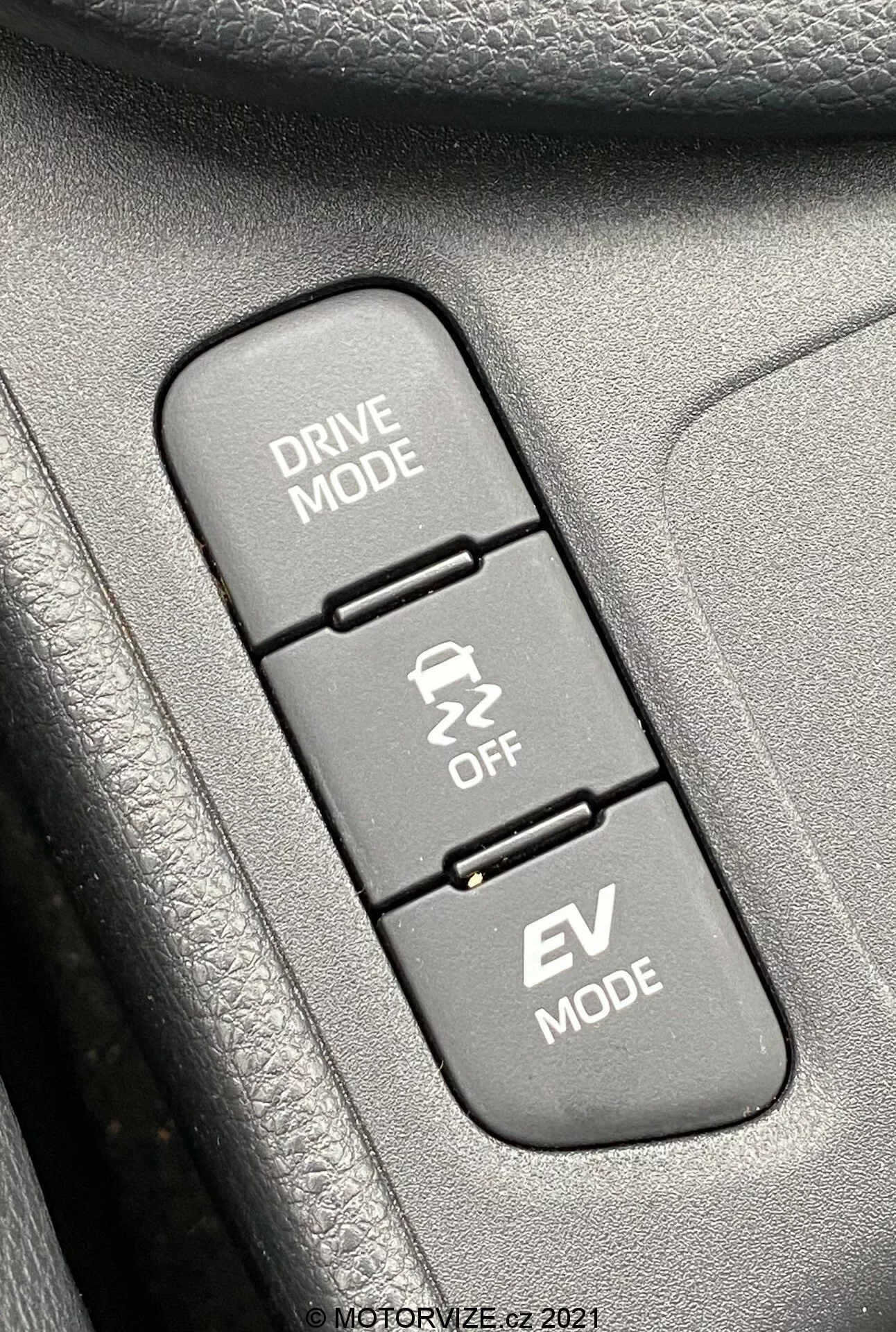Detailný záber interiéru Toyoty Yaris 2020, zameraný na tri vertikálne zarovnané ovládacie tlačidlá týkajúce sa režimov jazdy. Zhora nadol: 'DRIVE MODE' s piktogramom kľukatej cesty, 'Traction Control OFF' s piktogramom šmykľavého auta a 'EV MODE' indikujúceho funkciu elektrického vozidla. Tlačidlá sú namontované na textúrovanej ploche, pravdepodobne súčasťou dverového panela alebo stredovej konzoly. Obraz je ostro zaostrený na tlačidlá, pričom okolité textúry sú mierne rozmazané, rovnomerne osvetlené denným svetlom pre čistú viditeľnosť.