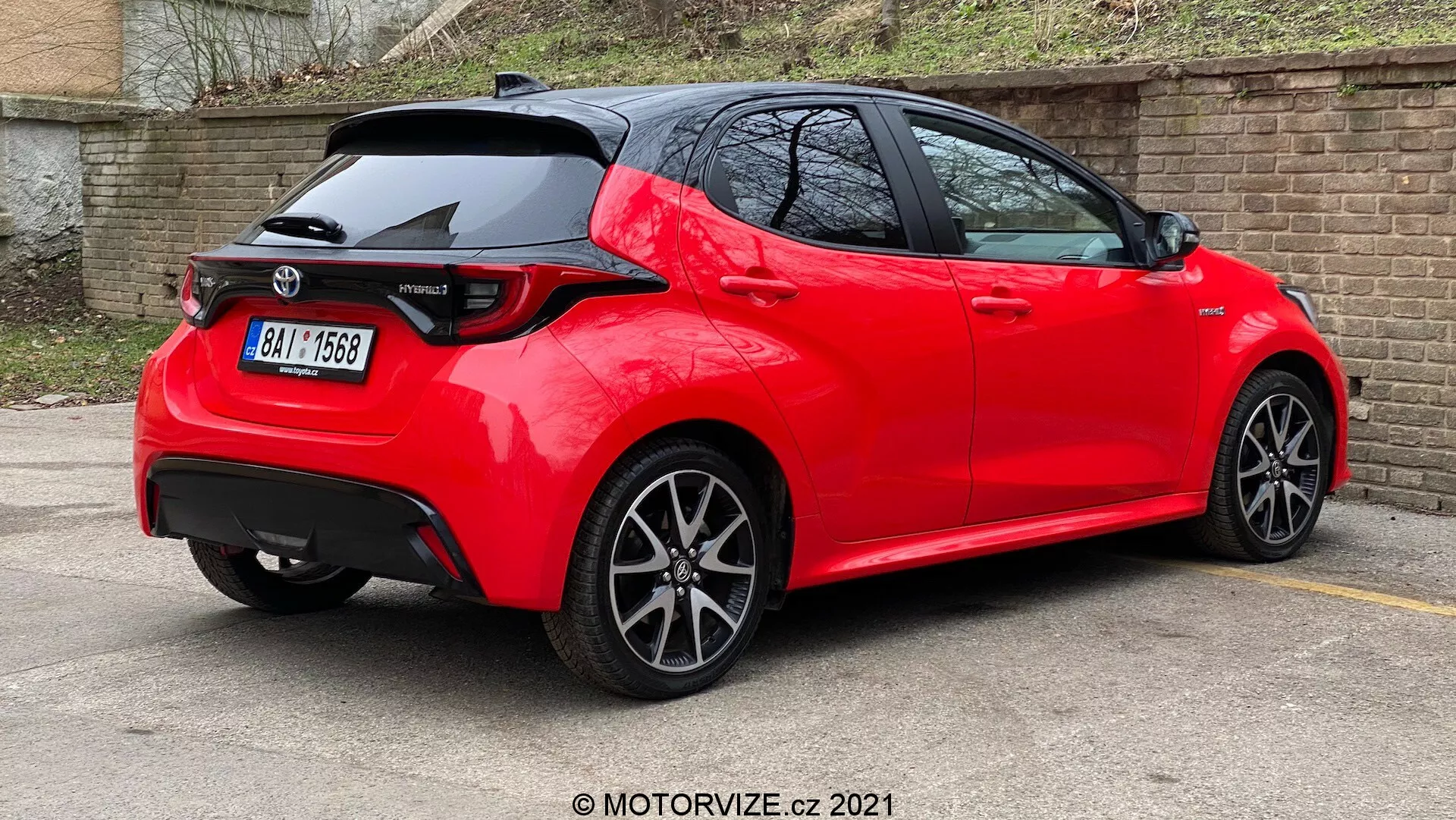 Trojtvrťový zadný pohľad na jasne červený, moderný hatchback s ostrými líniami a záhybmi, strešným spojlerom, zadnými svetlami typu LED, žraločou anténou, viaclúčovými zliatinovými kolesami a umiestnený na parkovisku s reflexným, dobre udržiavaným lakom, čo naznačuje nový alebo dobre udržiavaný model v športovej výbave.